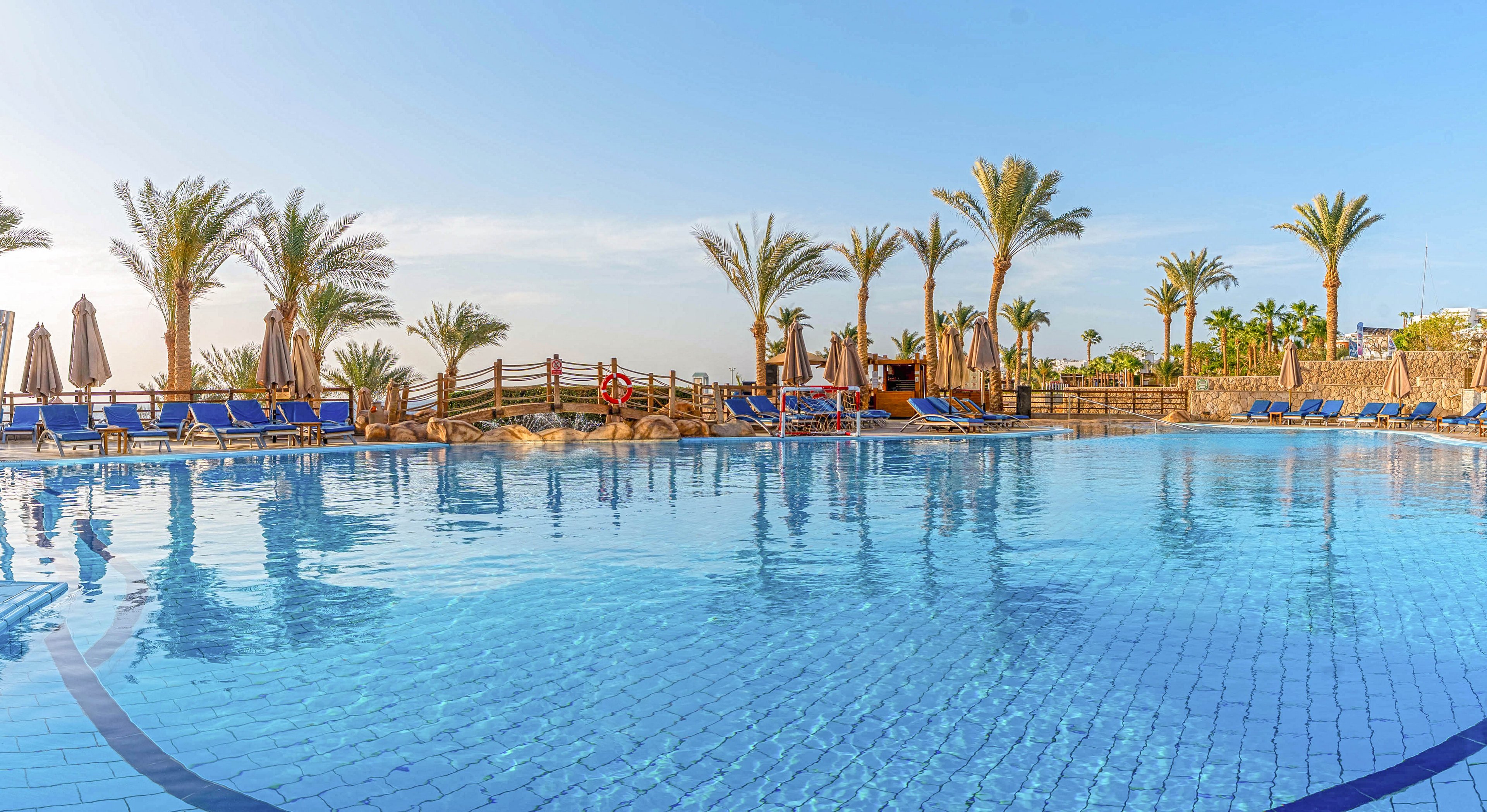 A large outdoor swimming pool reflects the clear blue sky at a sunny resort. Palm trees and sun loungers line the poolside, creating a relaxing vacation atmosphere. Umbrellas and a wooden fence border the area, with a lifebuoy visible near the edge. The scene suggests a tranquil holiday setting in a warm, coastal climate.