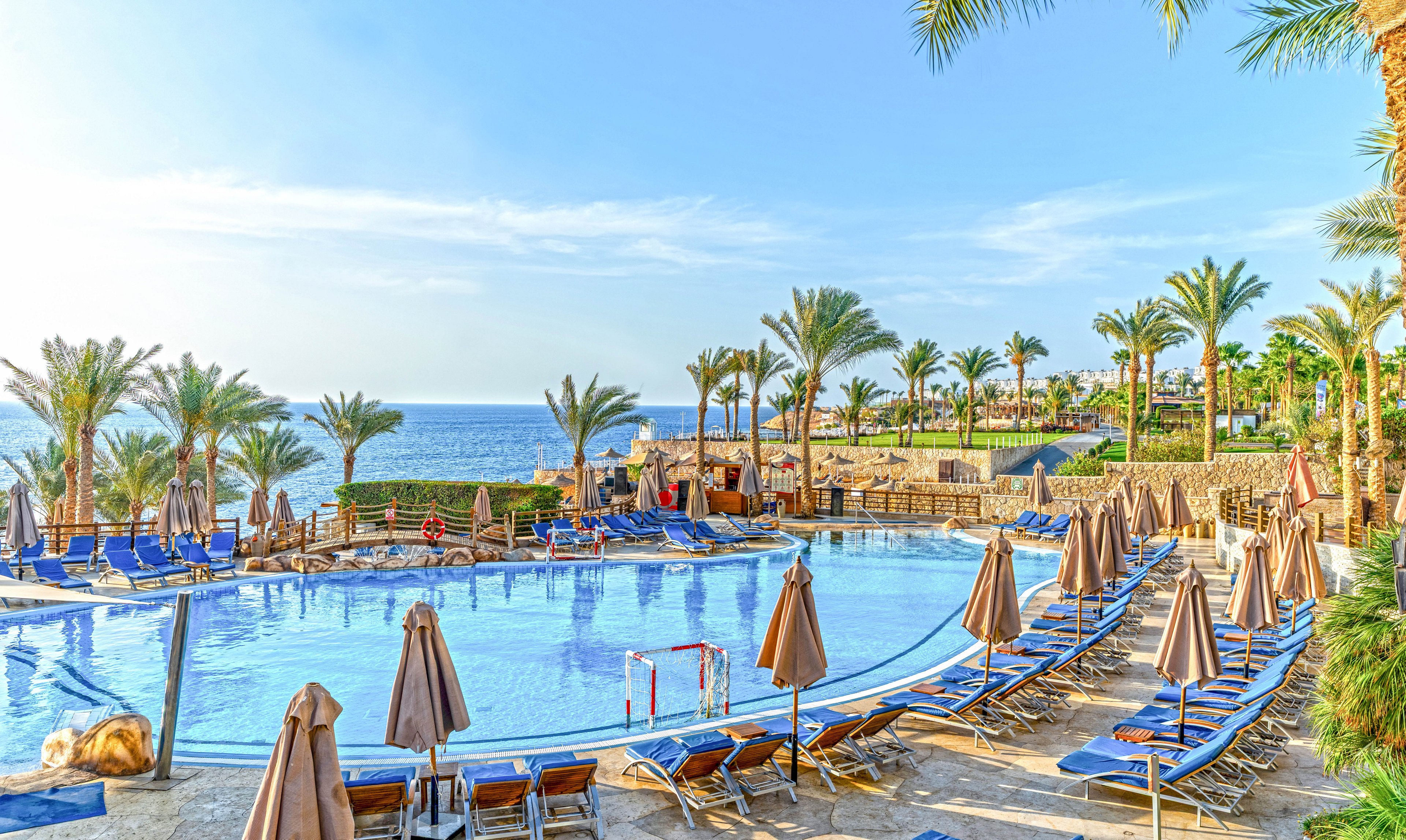 A large circular swimming pool is surrounded by rows of blue lounge chairs and beige umbrellas at a seaside resort. Tall palm trees frame the scene, with terraced stone walls and pathways leading toward the water. The calm sea is visible in the background under a clear blue sky. The bright sunlight and reflections on the pool create a warm, inviting vacation atmosphere.