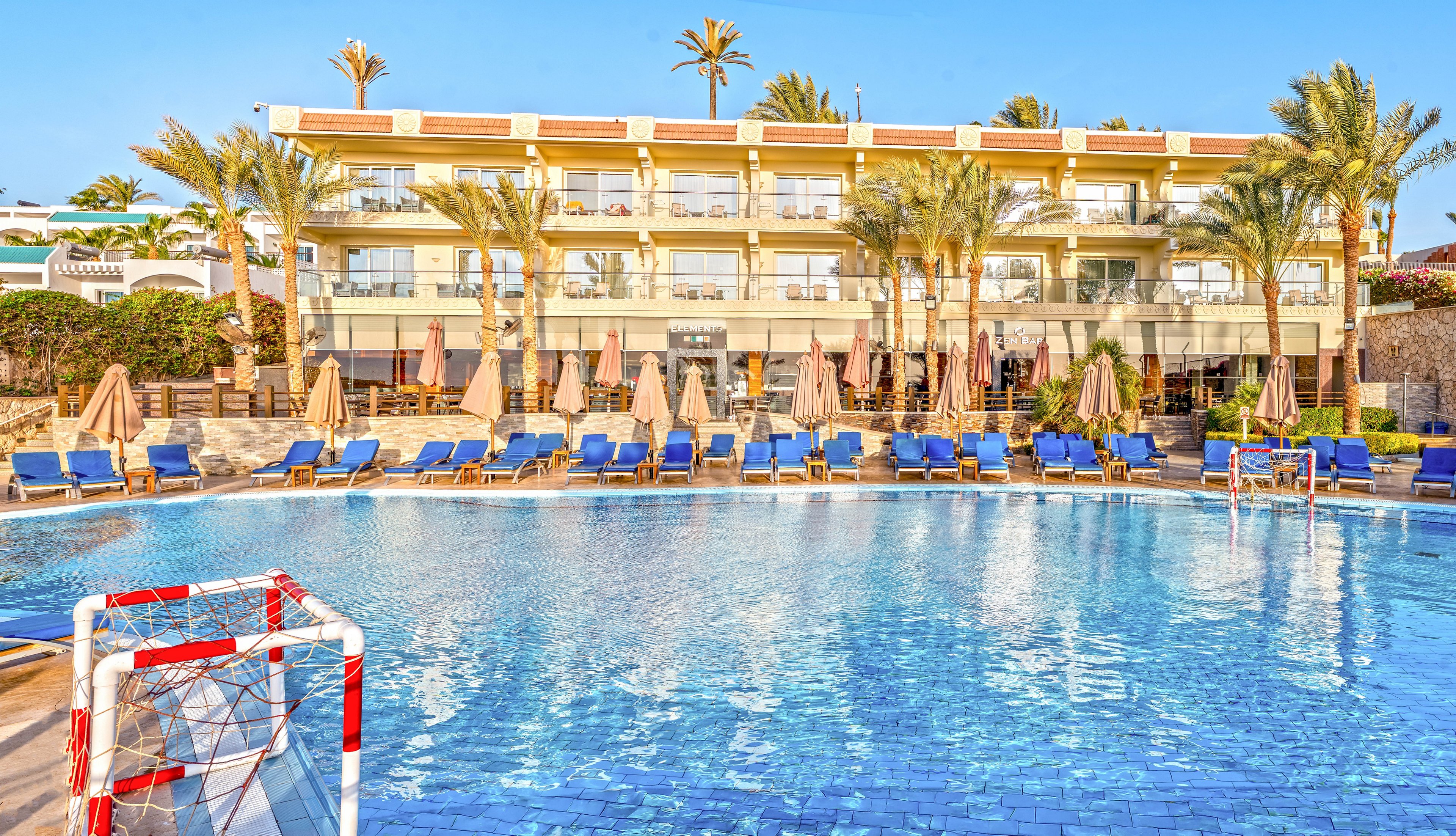 A large outdoor swimming pool dominates the foreground, reflecting the clear blue sky. Numerous blue sun loungers and beige umbrellas line the poolside, facing a multi-story hotel building with balconies. Palm trees rise above the structure, adding a tropical resort atmosphere. A small water polo or pool soccer goal is visible in the pool, suggesting recreational activities for guests.