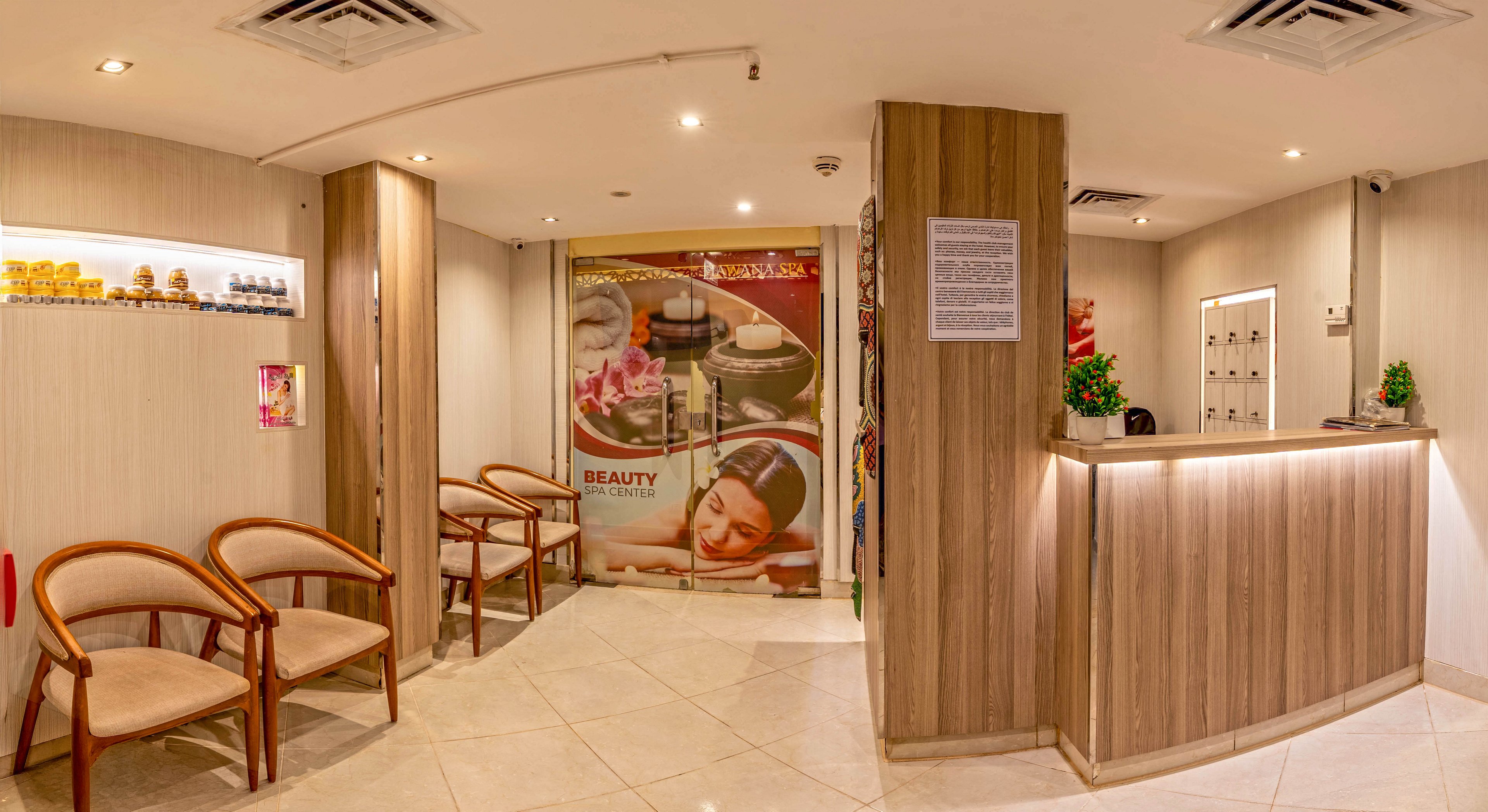 The image shows a contemporary beauty spa reception with a wooden front desk and soft lighting. A small waiting area with curved wooden chairs and cushioned seats is arranged along the left wall. Behind the reception, a large wall graphic features spa imagery including a woman receiving a facial and the word "BEAUTY" clearly visible. Shelves with small product jars and a decorative plant add detail to the calm, professional atmosphere. The overall palette is warm and neutral, emphasizing relaxation and comfort.