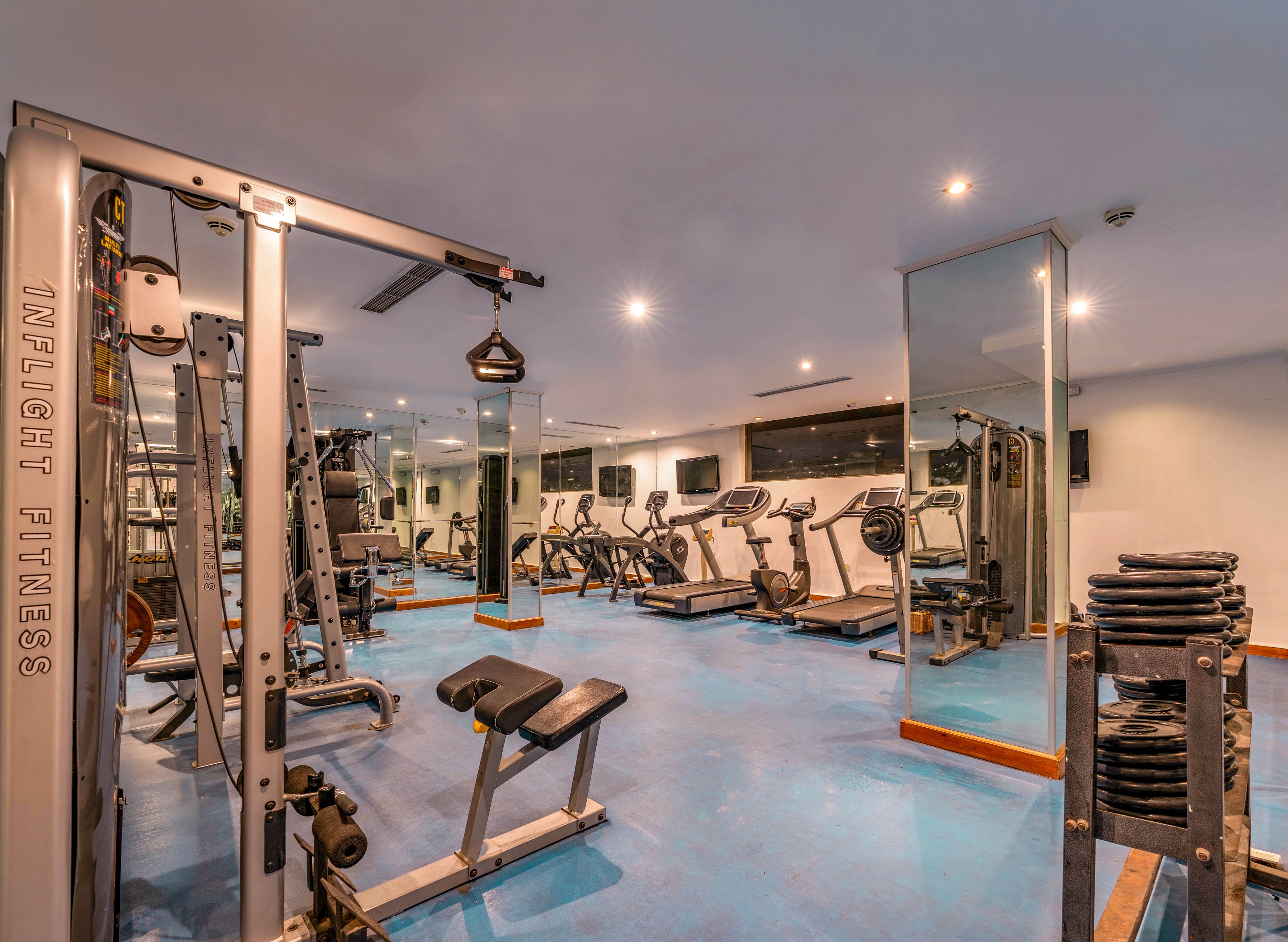 A modern indoor gym features a variety of strength and cardio equipment neatly arranged on a blue floor. Weight machines, benches, and free weights are positioned in the foreground, while treadmills and exercise bikes line the back wall. Large mirrors and bright ceiling lights create a spacious, reflective environment. The room appears clean, organized, and ready for individual workouts.