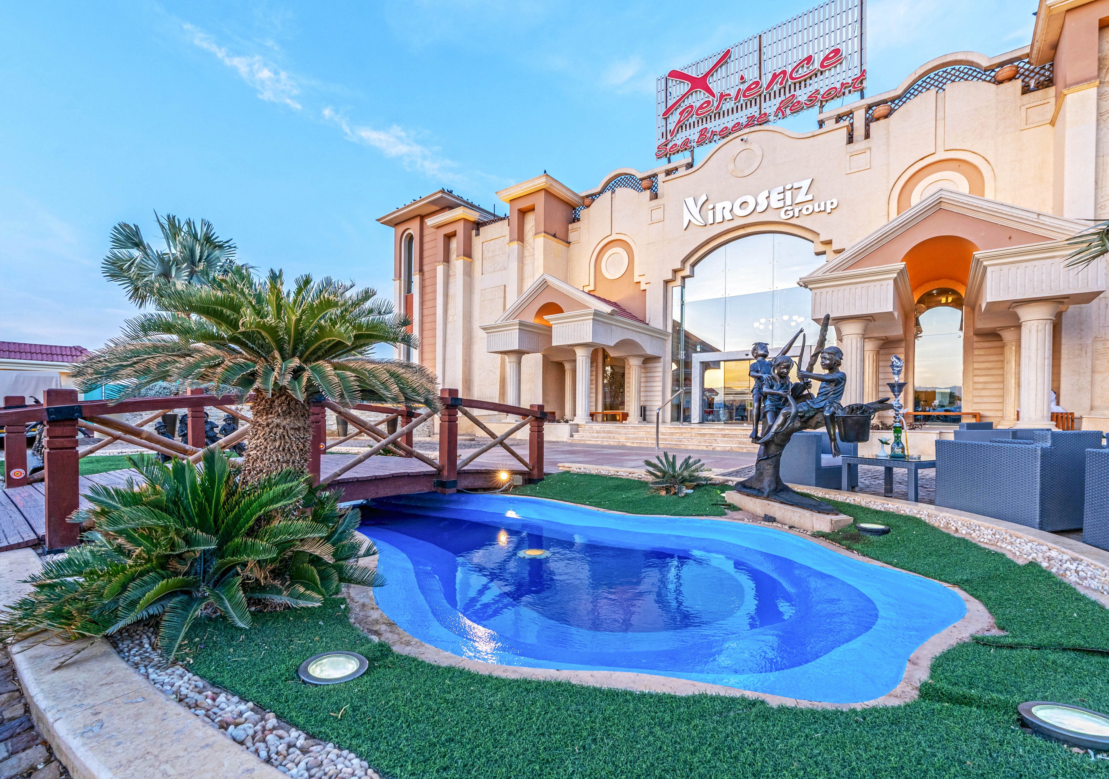 A luxurious resort entrance features a small, curved blue pool surrounded by artificial grass and landscaped stones. Palm trees and tropical plants frame a wooden bridge that crosses near the water. In the background, an ornate building façade displays large signage and architectural details in warm beige tones. A sculptural fountain with figures adds an artistic focal point to the outdoor area under a clear blue sky.