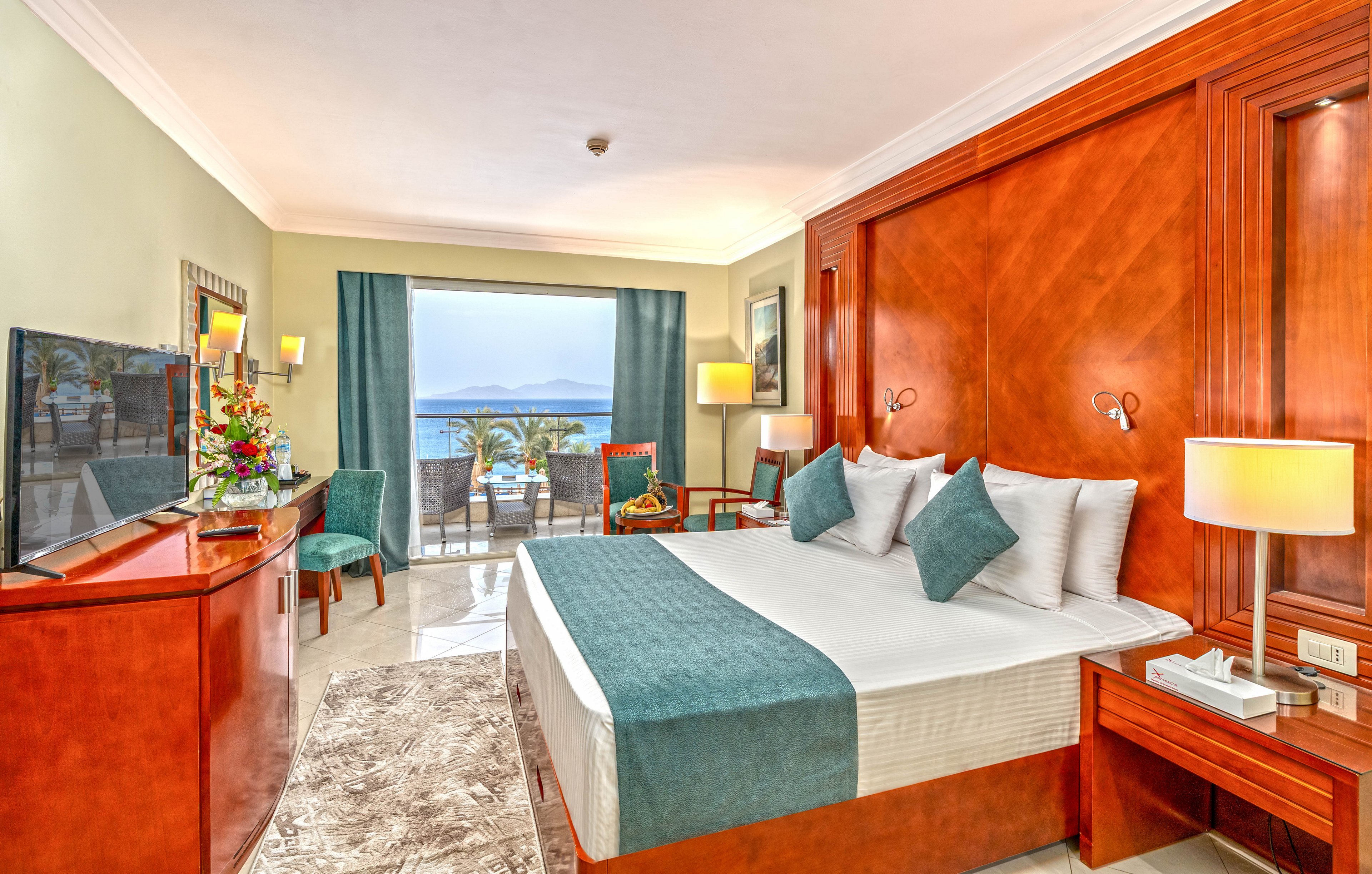 A spacious hotel room features a large bed with teal accents and multiple pillows. Warm wooden furniture, including a headboard, side tables, and a dresser with a flat-screen TV, frames the space. A sliding glass door opens to a balcony with chairs and a view of the ocean and distant mountains. Soft lighting from table lamps and natural daylight create an inviting, relaxing atmosphere.