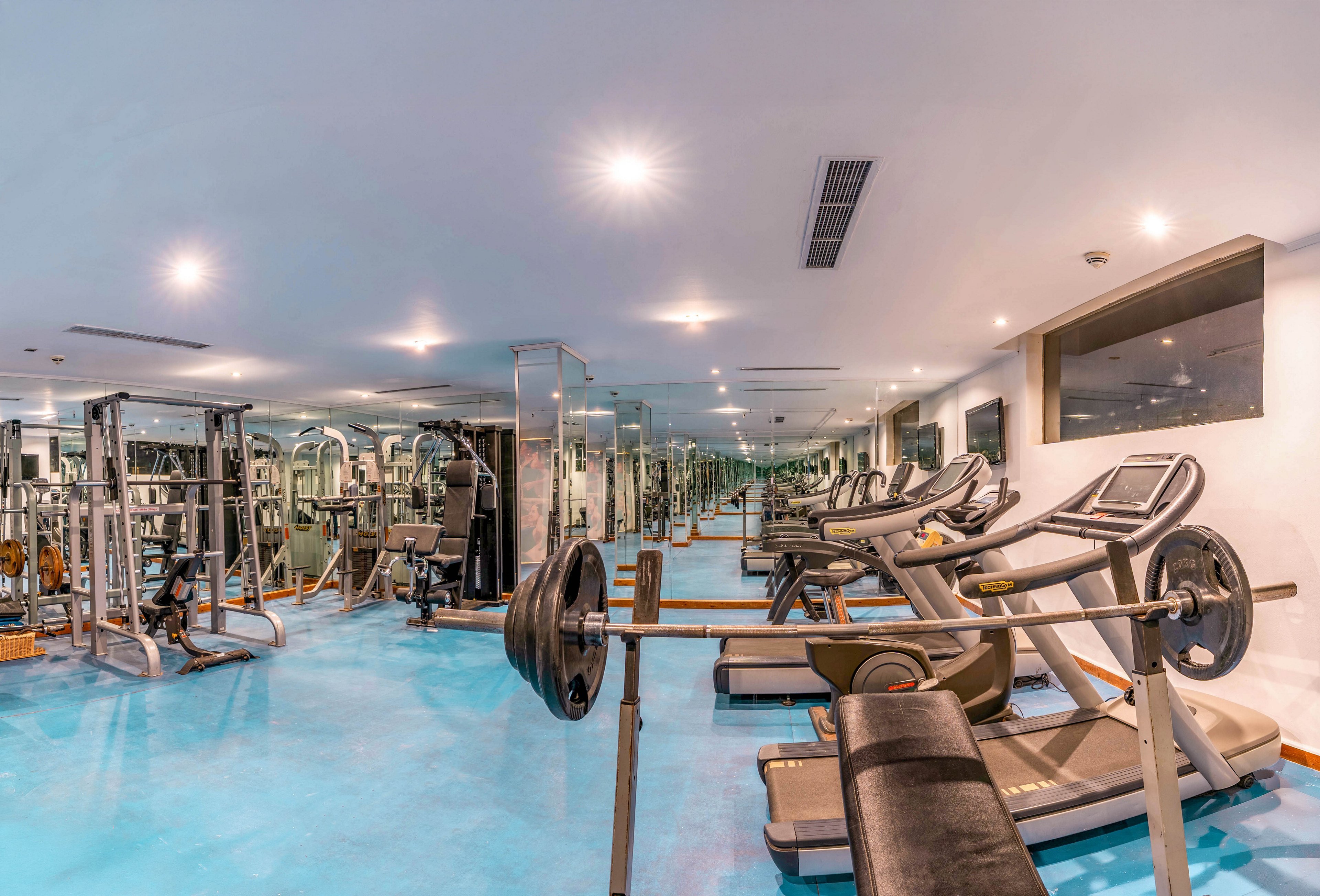 A spacious, modern gym interior features treadmills, weight benches, and strength-training machines arranged on a bright blue floor. Large mirrors line the walls, reflecting the equipment and creating a sense of depth. Overhead recessed lighting evenly illuminates the clean, organized workout space. The scene is empty of people, emphasizing the equipment and environment ready for exercise.