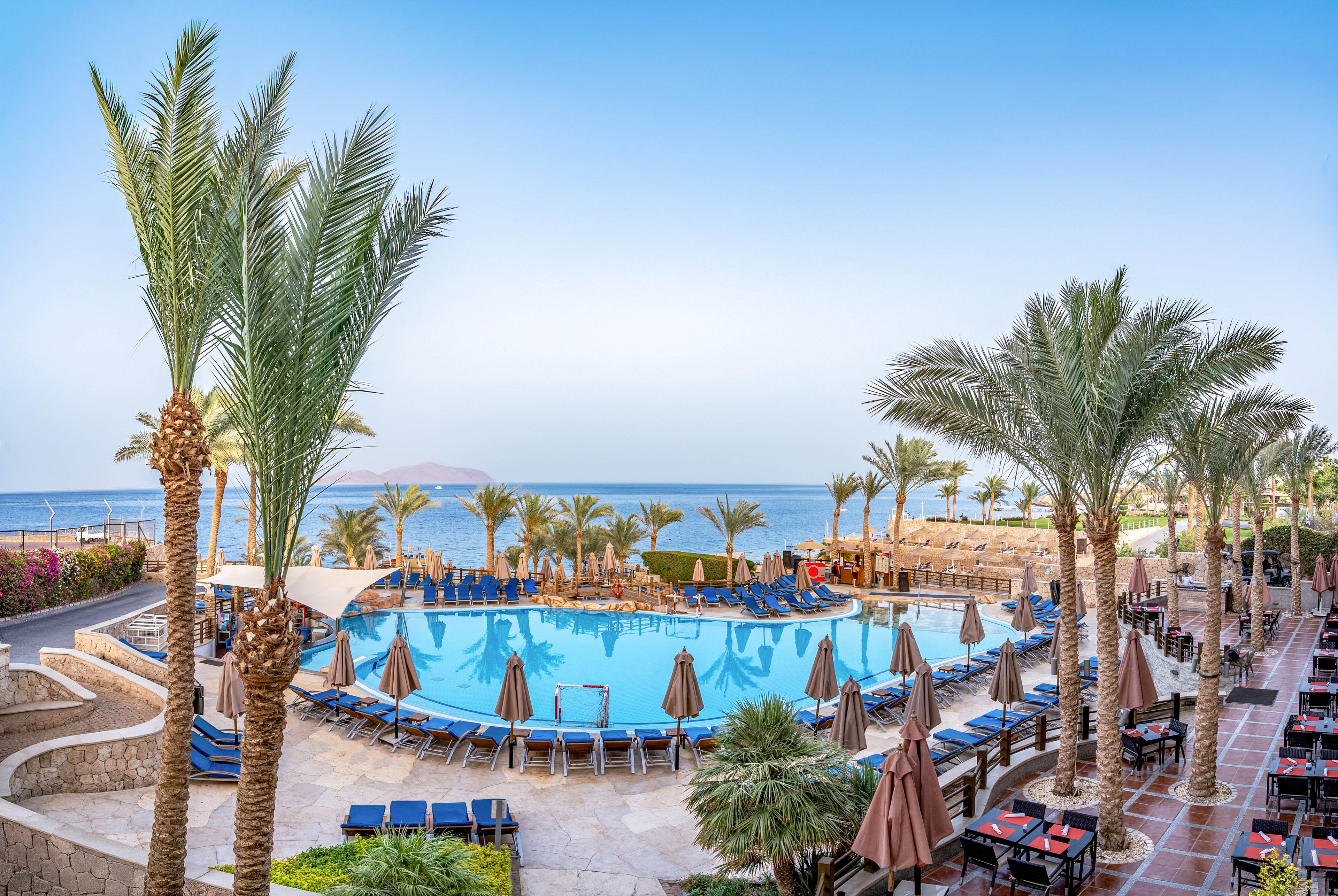 A large resort swimming pool is surrounded by sun loungers and umbrellas on a multi-level terrace. Tall palm trees frame the scene and lead the eye toward the calm blue sea in the background. The sky is clear, creating a bright and inviting vacation atmosphere. Tables and chairs line the walkway, suggesting an outdoor dining or relaxation area near the water.