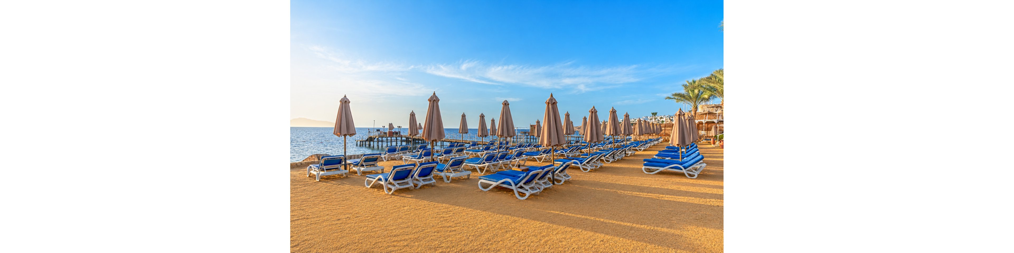 A wide sandy beach is lined with rows of blue sun loungers and closed beige umbrellas facing the sea. The scene appears calm and empty, suggesting early morning or off-season at a seaside resort. A wooden pier extends into the water, with distant landforms visible on the horizon. Clear blue sky and gentle sunlight create a warm, inviting coastal atmosphere.