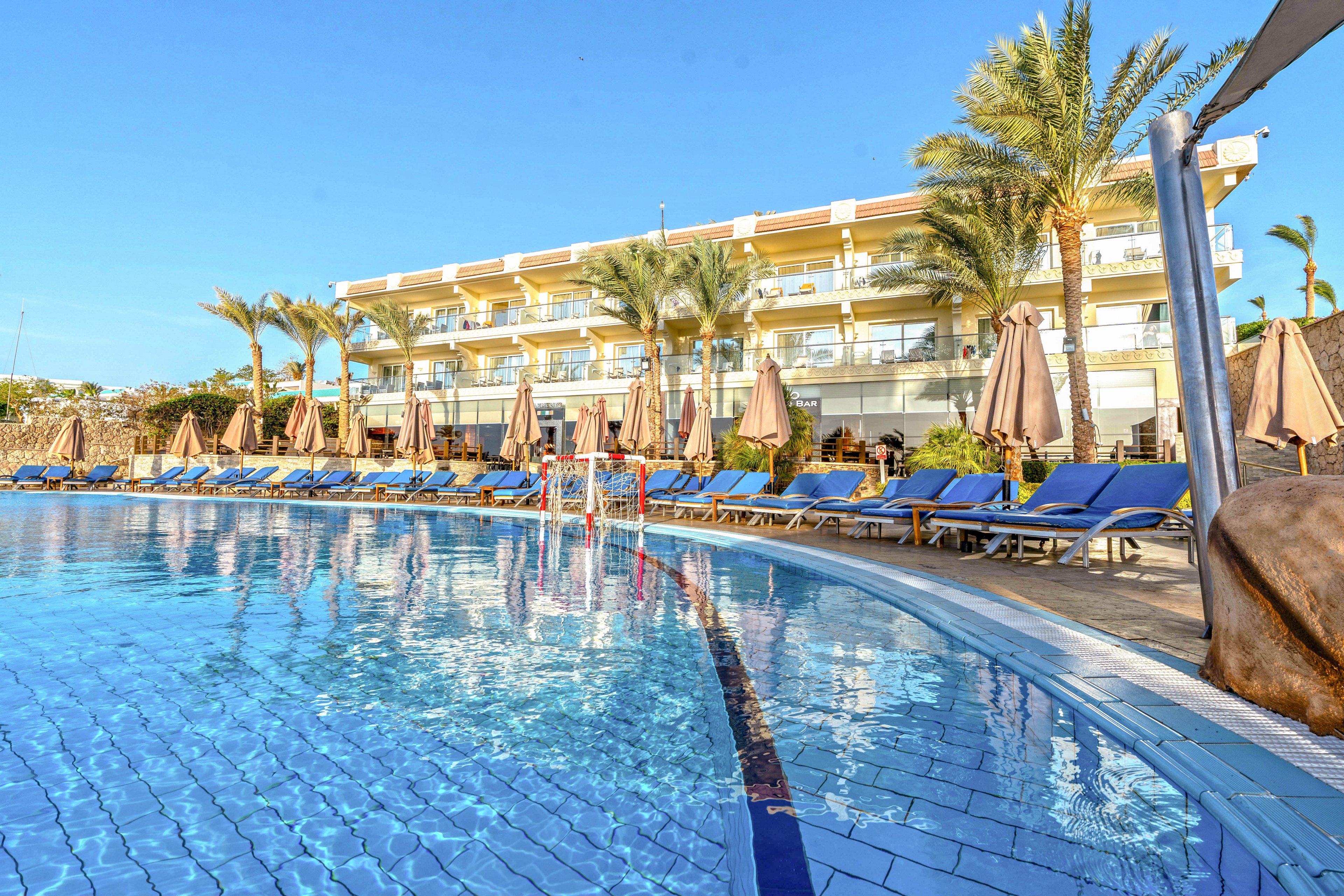 A large outdoor swimming pool reflects the clear blue sky in front of a modern resort building. Rows of blue sun loungers and closed beige umbrellas line the poolside, suggesting a relaxing vacation setting. Palm trees frame the scene, adding a tropical atmosphere. The bright sunlight and calm water create a warm, inviting holiday mood.