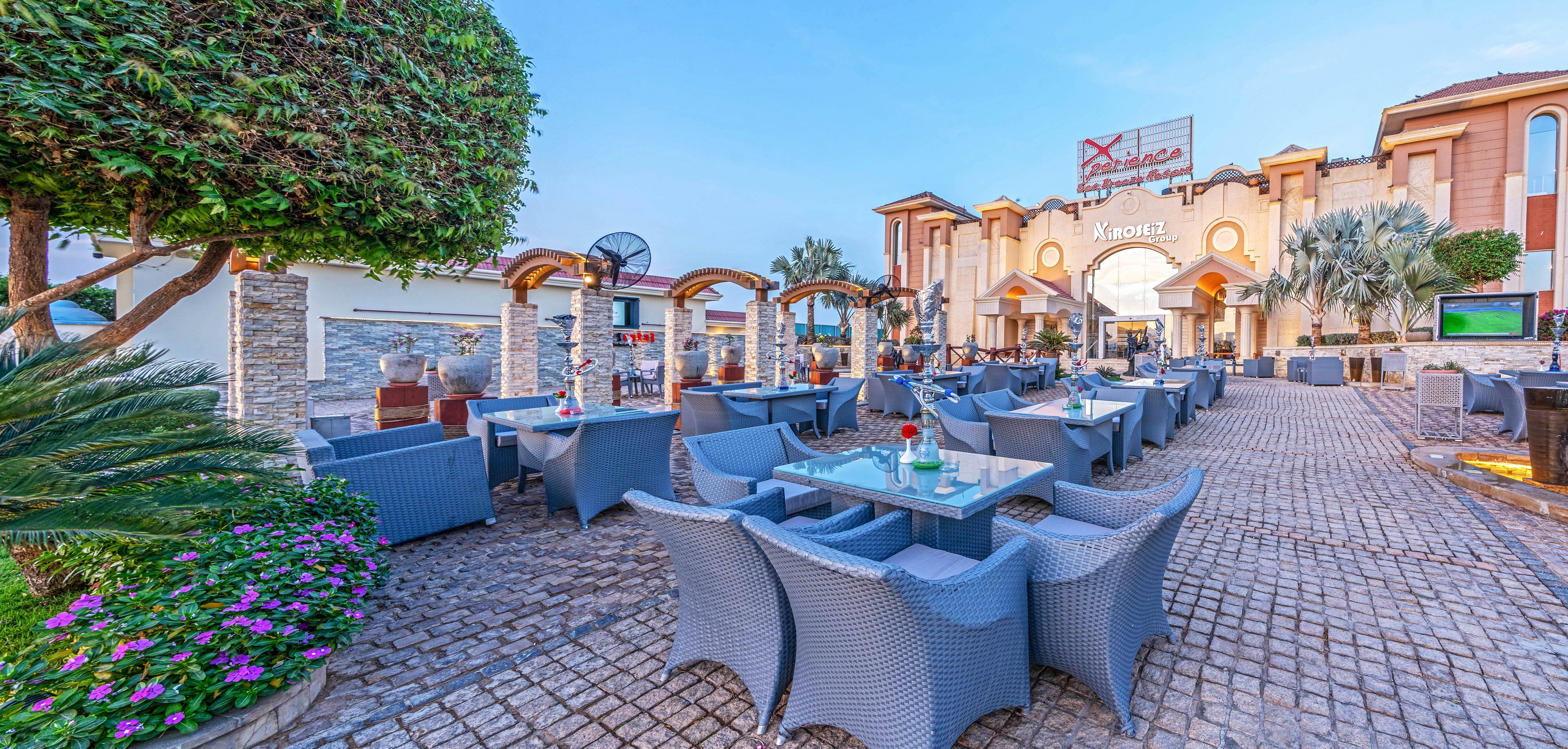 An expansive outdoor terrace at a resort features rows of gray wicker chairs and glass-top tables arranged on a cobblestone patio. The setting is framed by manicured trees, flowering plants, and stone columns with decorative arches. In the background, a large building with warm-toned facades and palm trees creates a welcoming entrance. The clear blue sky and bright daylight give the scene a relaxed, vacation atmosphere.