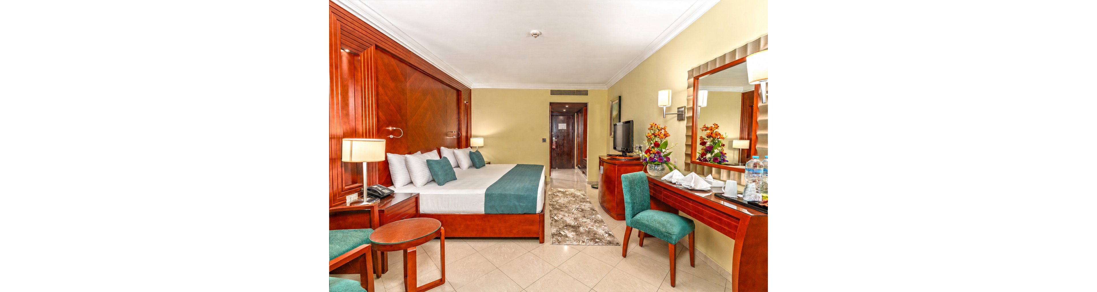 A spacious hotel room features a large bed with white linens and teal cushions, framed by rich wooden furniture. The room includes a seating area with a round table, upholstered chairs, and a long desk holding a flat-screen television and decorative flower arrangements. Warm wall sconces and table lamps create a bright, inviting atmosphere, while a hallway leads toward the entrance door. A neutral tiled floor and a soft rug add comfort and contrast to the polished wood and teal color palette.