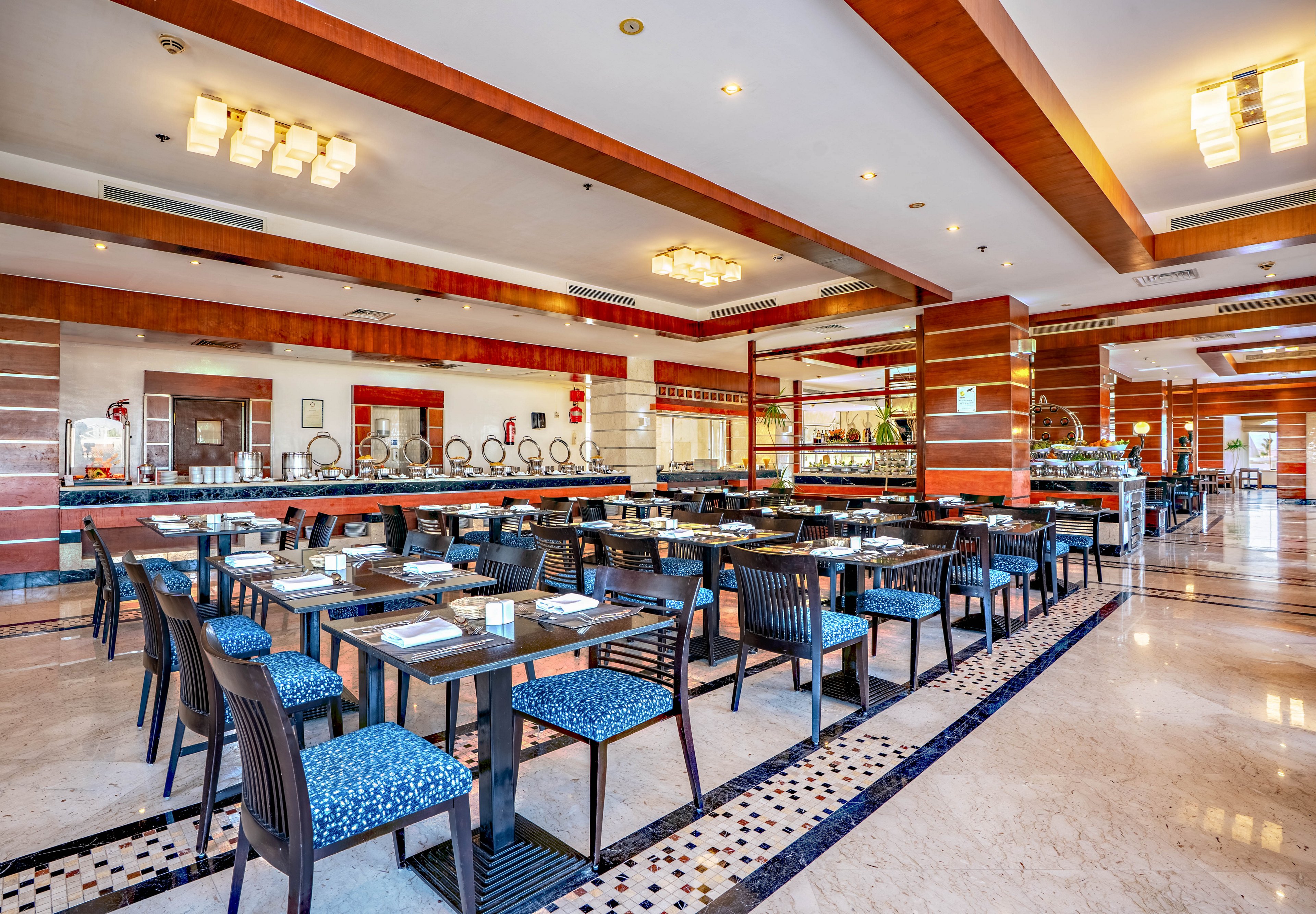 A spacious modern restaurant dining area is shown with neatly arranged tables and chairs. The room features polished marble floors, wooden ceiling beams, and contemporary light fixtures. Place settings are prepared on each table, and a buffet counter with covered dishes is visible in the background. Large windows and bright lighting create an inviting and clean atmosphere suitable for hotel or resort dining.