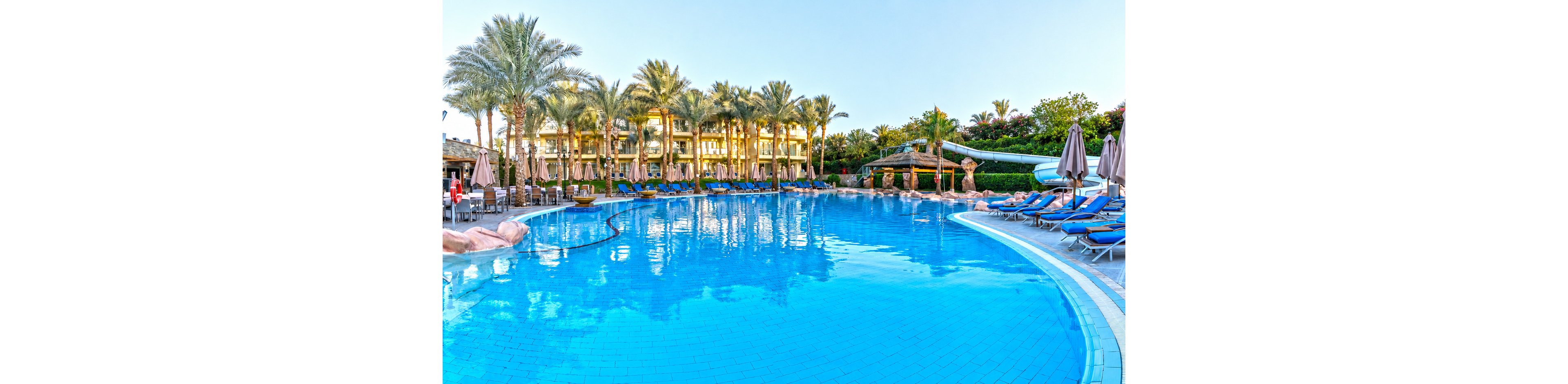 A large, clear blue resort swimming pool curves across the foreground, reflecting the surrounding palm trees and bright sky. Sun loungers with umbrellas line the poolside, suggesting a relaxing vacation setting. In the background, a multi-story hotel building and lush greenery frame the scene. The calm water and soft lighting create a tranquil, inviting atmosphere for guests.
