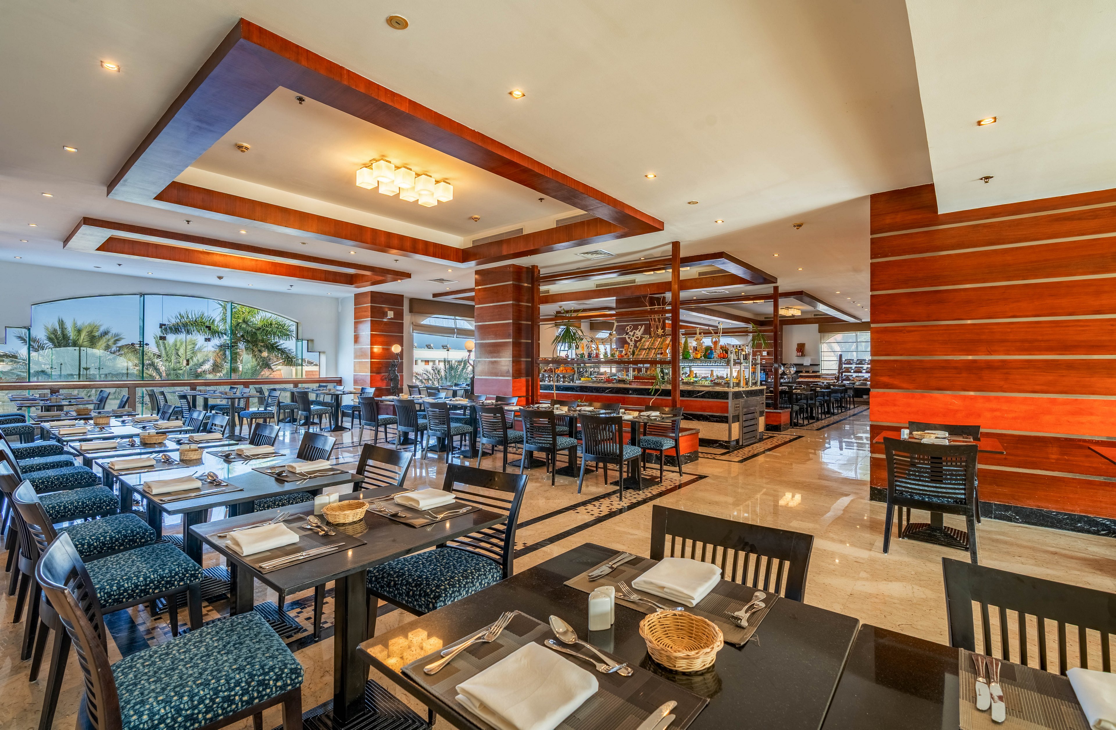 A spacious, modern restaurant dining room is shown with neatly arranged tables and chairs. Each table is set with cutlery, napkins, glasses, and small decorative baskets, ready for guests. Warm wooden paneling, ceiling details, and soft lighting create an inviting atmosphere. Large windows on one side reveal greenery and natural light, enhancing the open, airy feel of the space.
