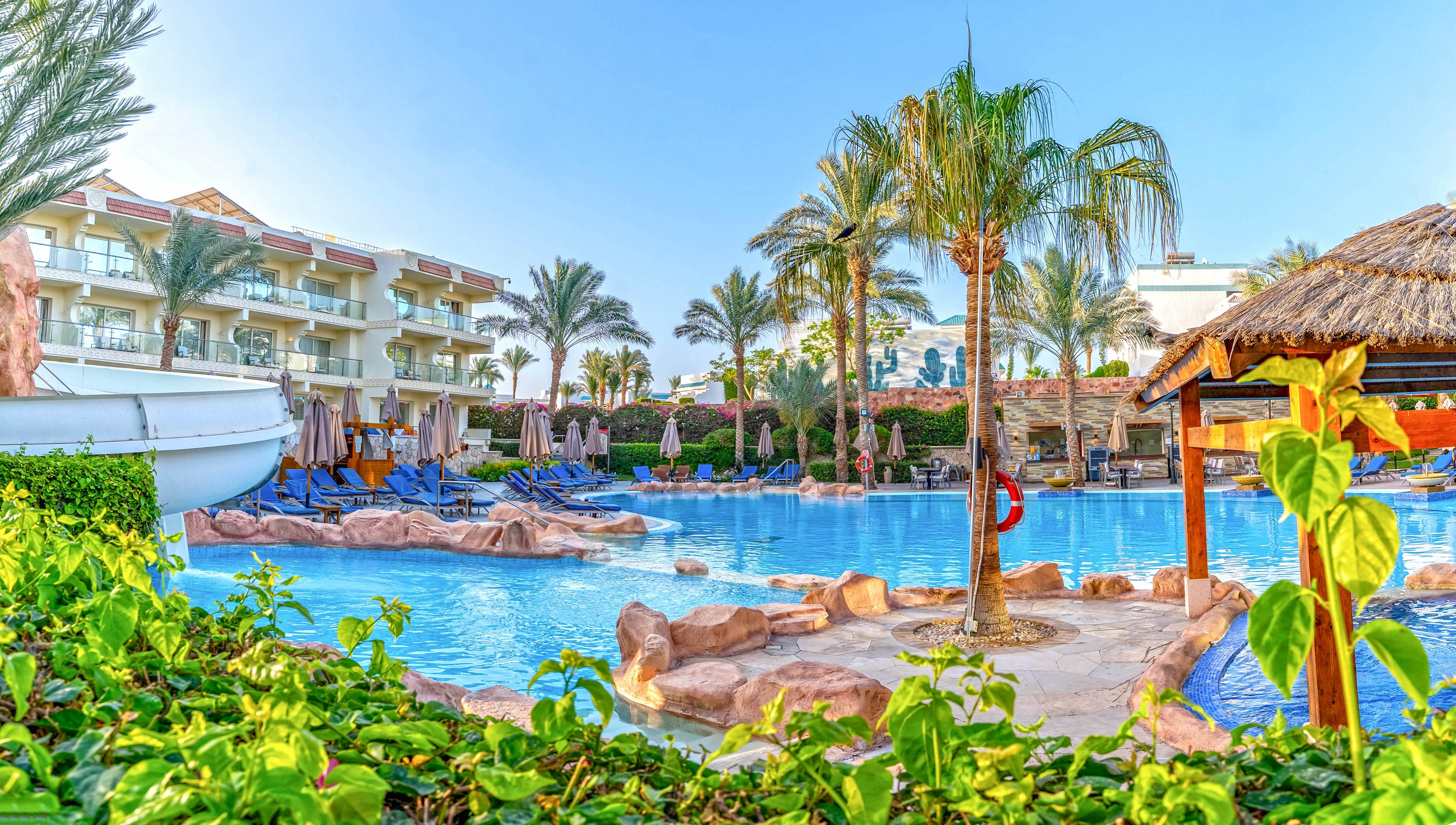 A large resort-style swimming pool is surrounded by palm trees and lush greenery. Sun loungers and umbrellas line the poolside near multi-story hotel buildings. Rock formations and a thatched-roof structure add a tropical, vacation atmosphere. The clear blue water and bright sky suggest a warm, sunny day at a holiday destination.