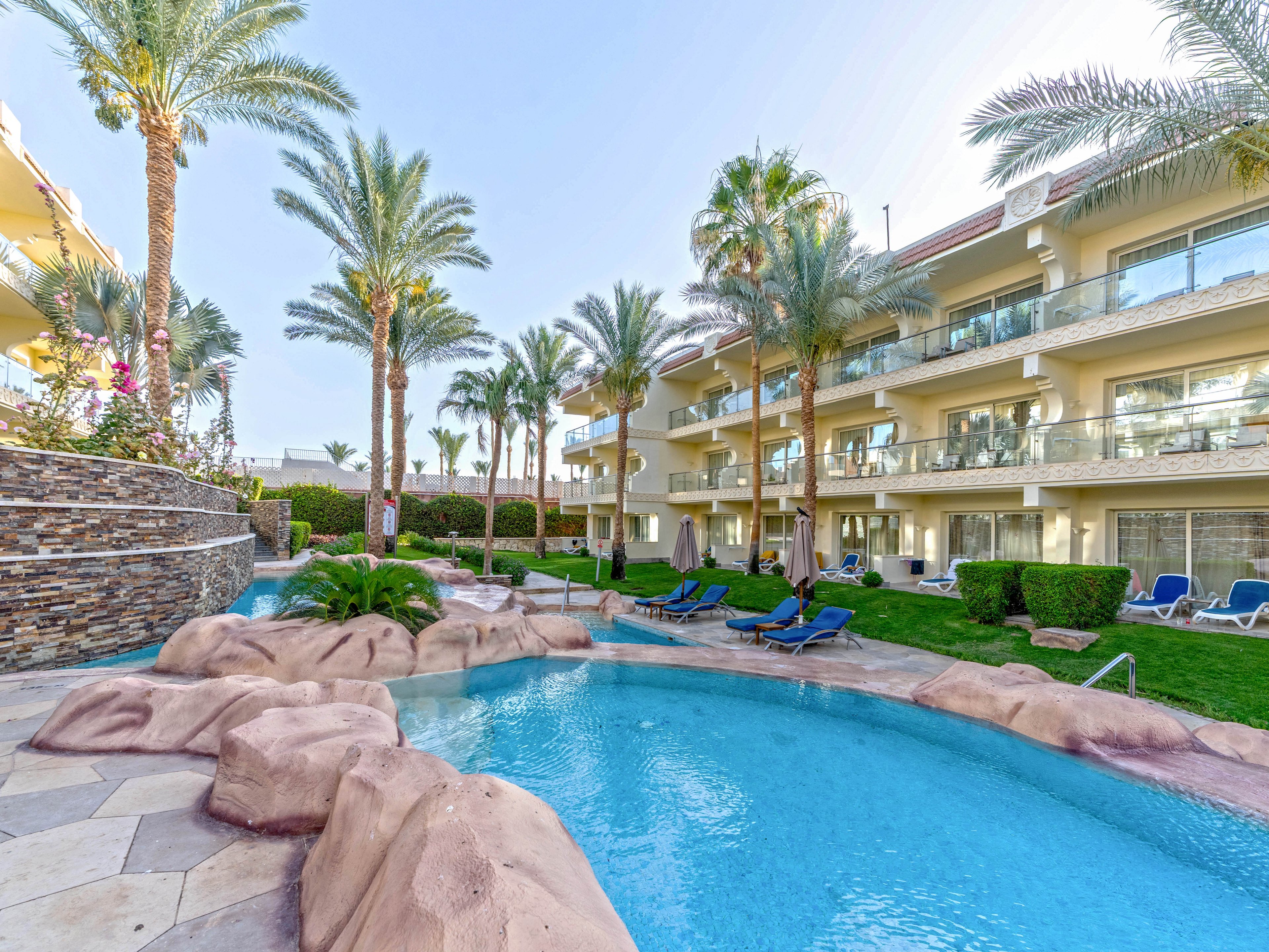 A resort-style swimming pool winds through a landscaped courtyard surrounded by palm trees. The pool features curved edges, rock formations, and clear blue water, creating a tropical oasis atmosphere. Multi-story hotel or apartment buildings with balconies overlook the pool area. Sun loungers and neatly trimmed hedges add to the relaxing vacation setting.
