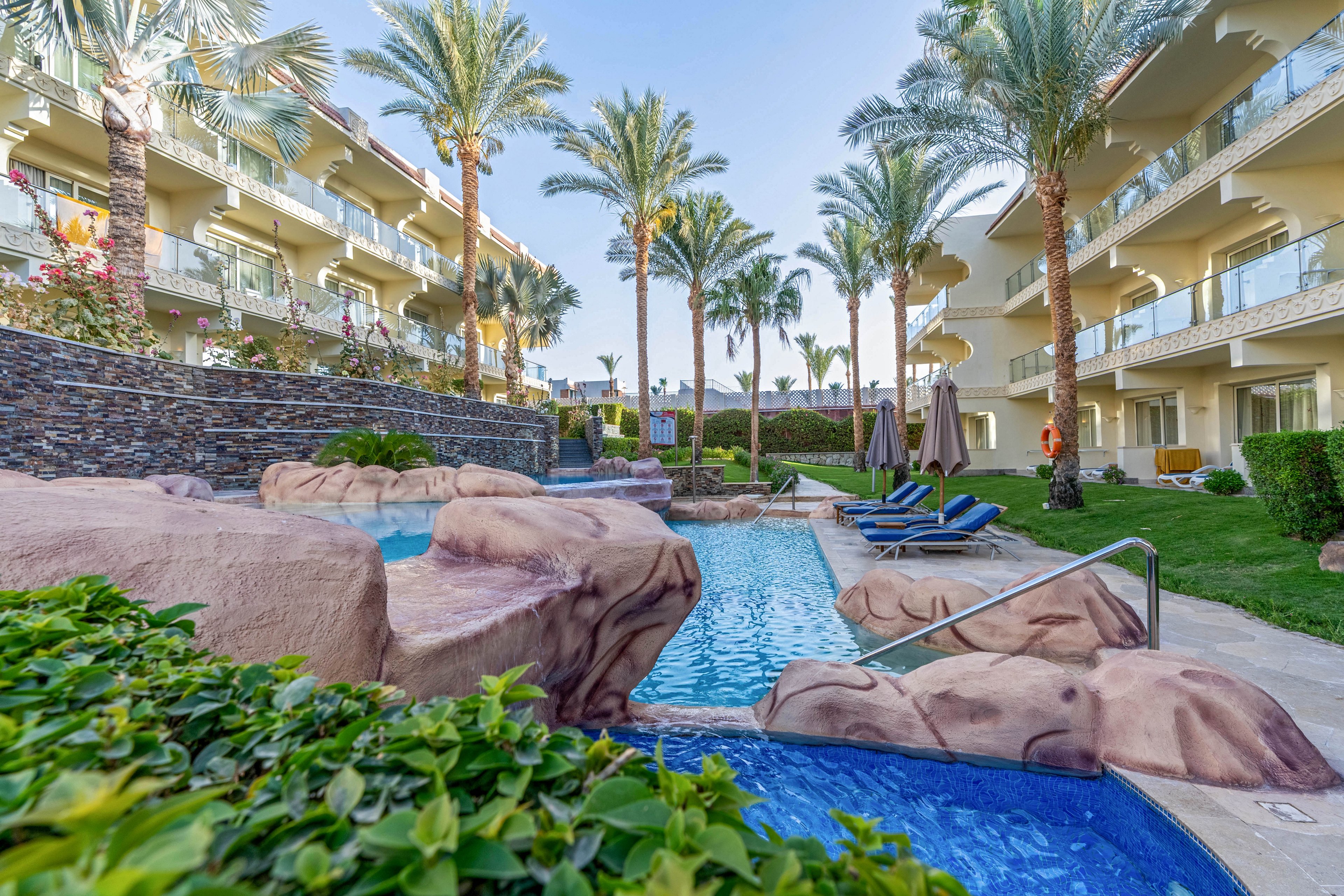 A resort-style swimming pool winds through a landscaped courtyard between multi-story hotel buildings. Artificial rock formations frame the pool, with stainless steel handrails leading into the water. Several blue lounge chairs and umbrellas are arranged along the poolside, suggesting a relaxing vacation setting. Tall palm trees and manicured greenery add a tropical atmosphere under a clear blue sky.