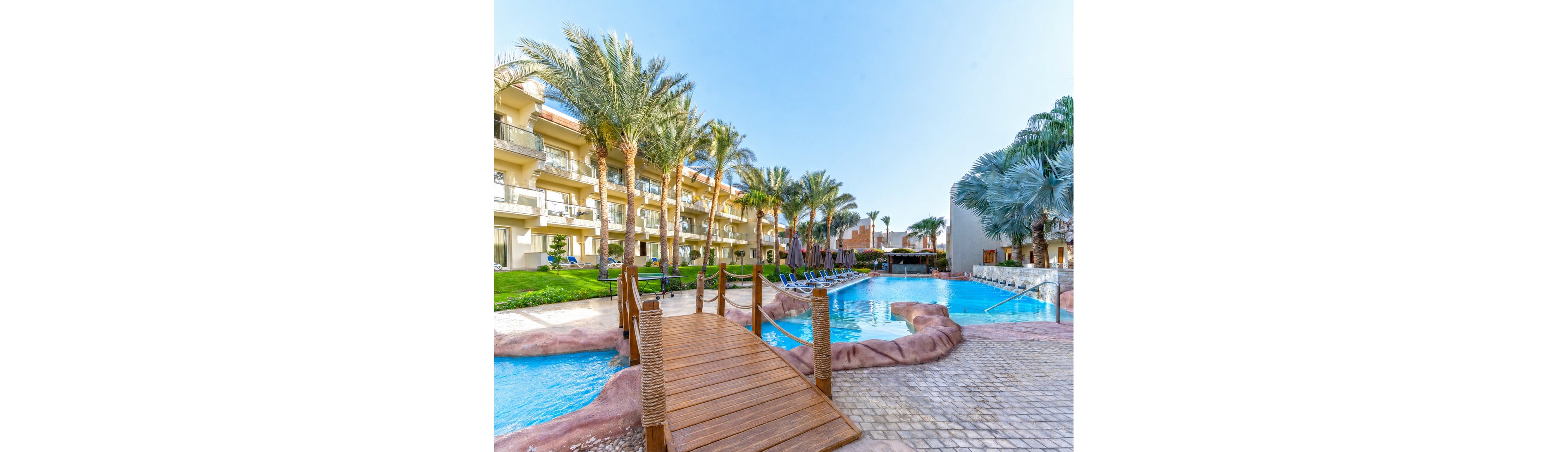 A resort-style swimming pool is surrounded by tall palm trees and a multi-story hotel building with balconies. A wooden footbridge crosses over the bright blue water, connecting stone pathways around the pool. Sun loungers are arranged along the poolside, suggesting a relaxing vacation setting. The scene is brightly lit under a clear blue sky, emphasizing a warm and tropical atmosphere.