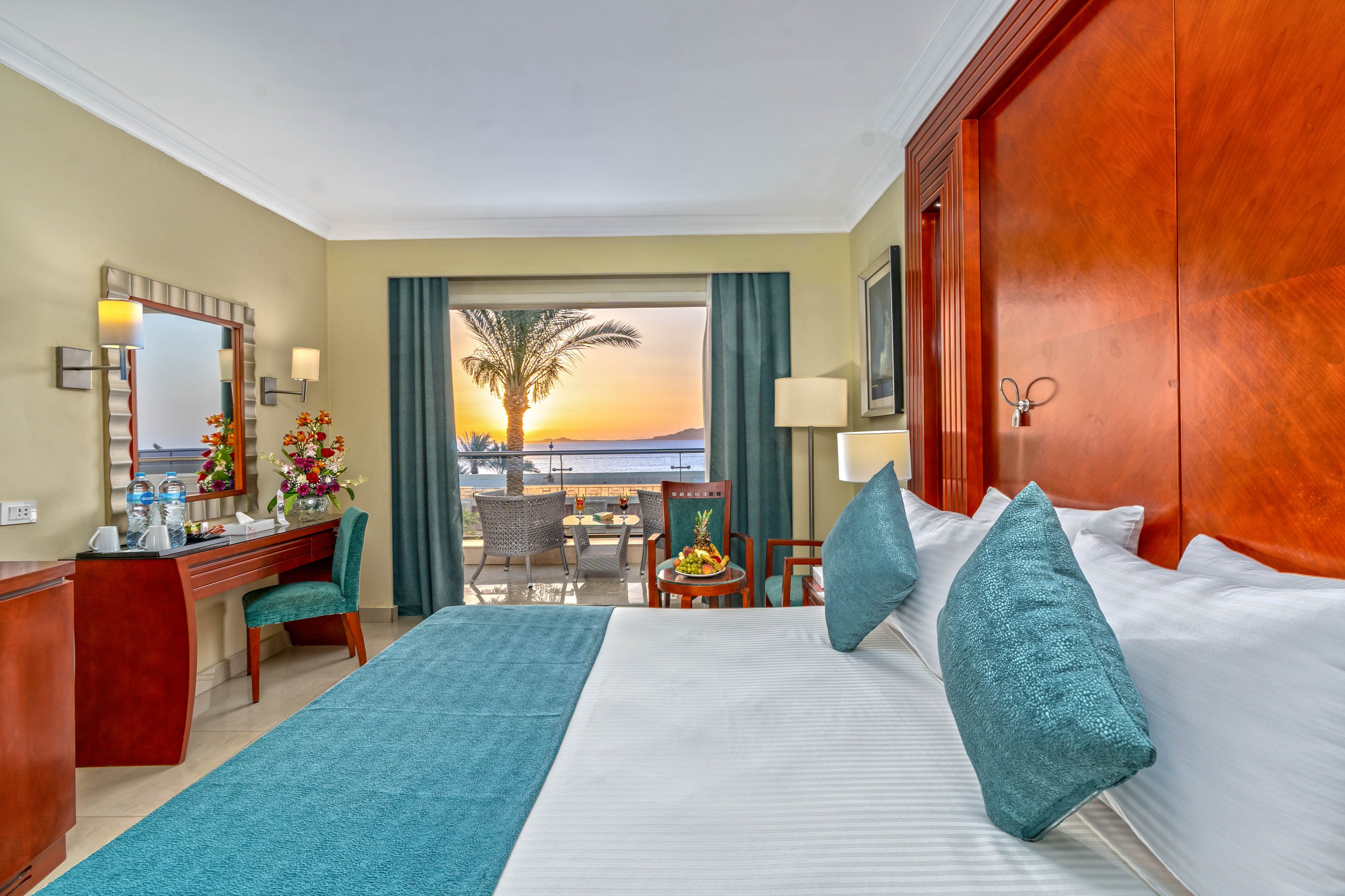 A spacious hotel room features a large bed with teal accents and wooden furnishings. The room opens onto a balcony overlooking the ocean at sunset, with a palm tree framing the view. A desk with a mirror, chair, and decorative flowers sits to the left, while a small table with fruit and drinks is arranged on the balcony. Soft lighting and coordinated textiles create a calm, inviting atmosphere ideal for vacation or resort stays.