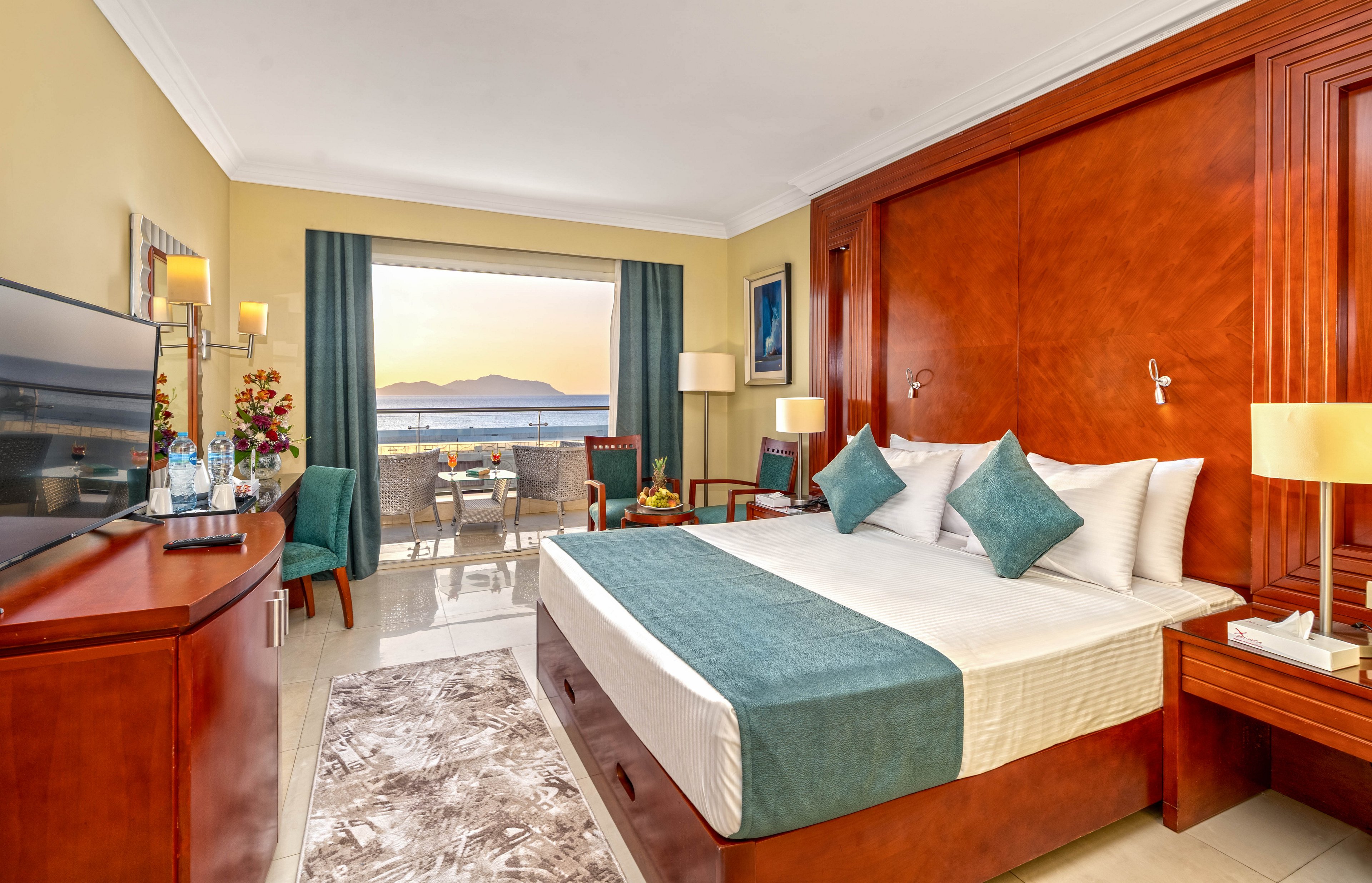 A contemporary hotel room features a large bed with teal accents and wooden furniture. The space opens onto a balcony with chairs and a table overlooking the ocean and distant mountains at sunset. Soft lighting from bedside lamps and a floor lamp creates a warm, inviting atmosphere. A flat-screen TV, decorative flowers, and neatly arranged amenities complete the comfortable interior.