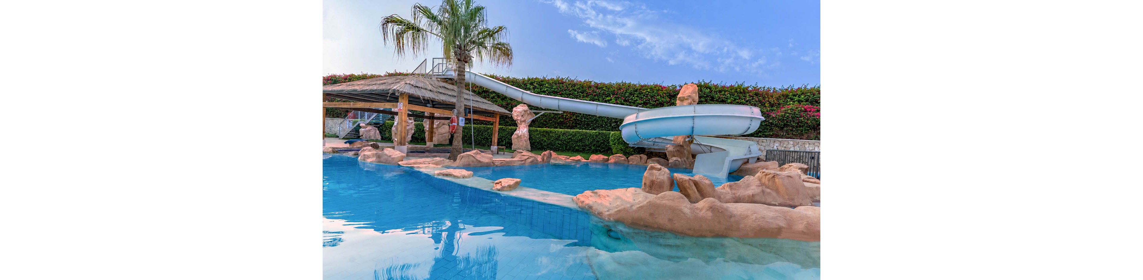 An outdoor resort-style swimming pool features a large curved waterslide ending in clear blue water. Artificial rock formations and a thatched-roof structure frame the pool area, creating a tropical atmosphere. A tall palm tree and manicured hedges line the background under a bright blue sky with light clouds. The scene is calm and empty, suggesting a relaxing leisure setting.