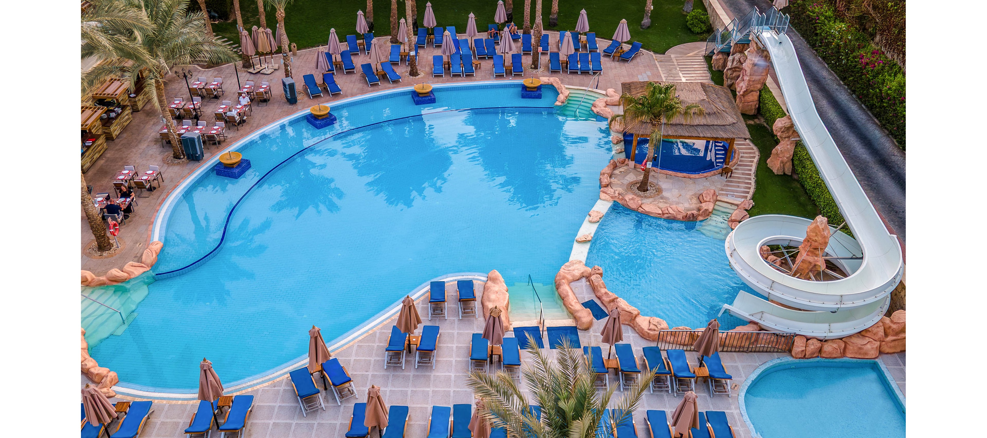 A large resort-style swimming pool is surrounded by numerous blue sun loungers and beige umbrellas. A curved white waterslide leads into a smaller adjoining pool area framed by artificial rock formations. Palm trees and landscaped greenery border the pool deck, adding a tropical atmosphere. Tables and chairs are visible near the top of the image, suggesting an outdoor dining or bar area.