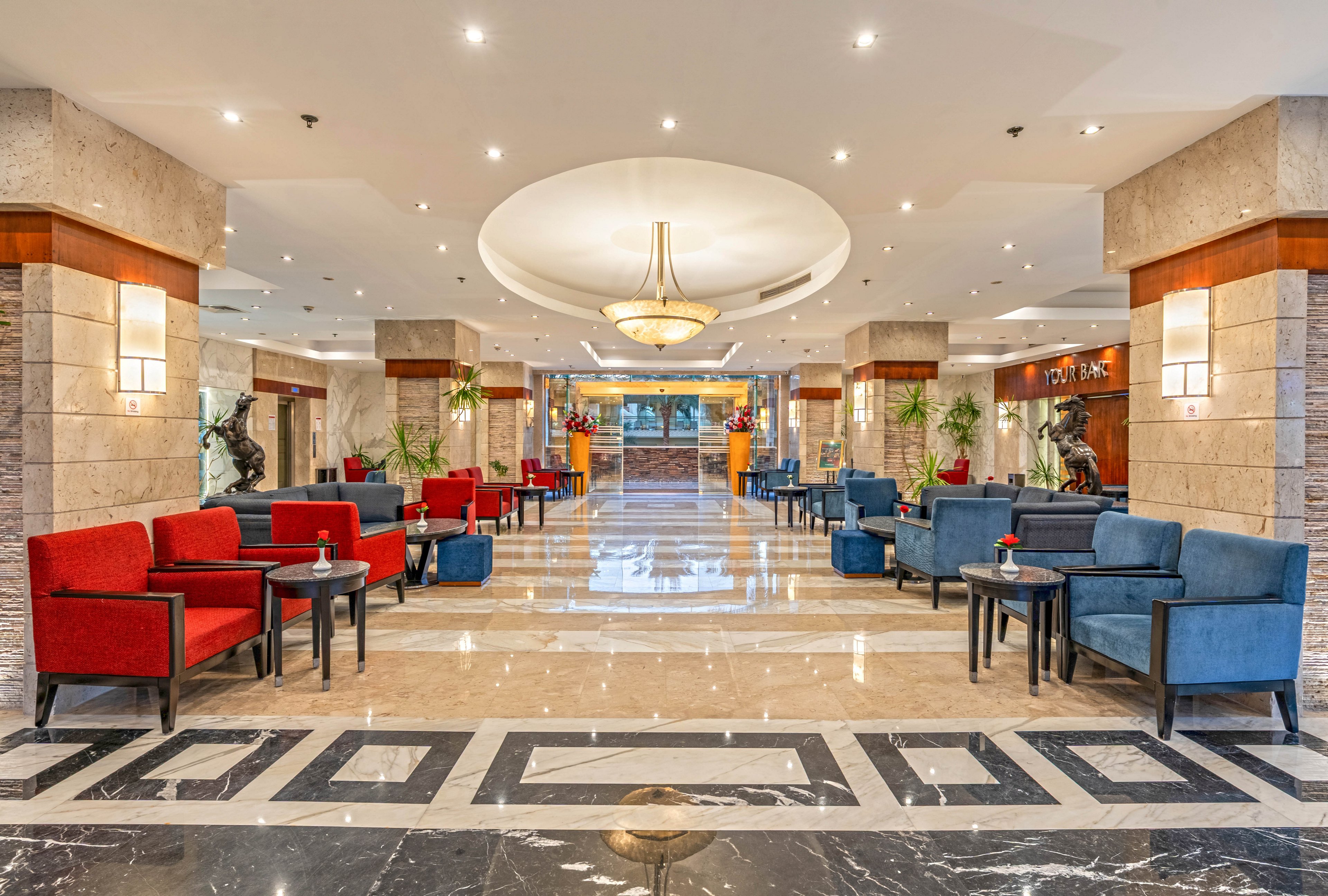 A large, modern hotel lobby features rows of red and blue armchairs arranged around small side tables. The polished marble floor and decorative black-and-white border lead the eye toward a central chandelier and reception area in the background. Warm ceiling lights and neutral stone walls create a bright, welcoming atmosphere. Potted plants and sculptures add decorative accents throughout the space.