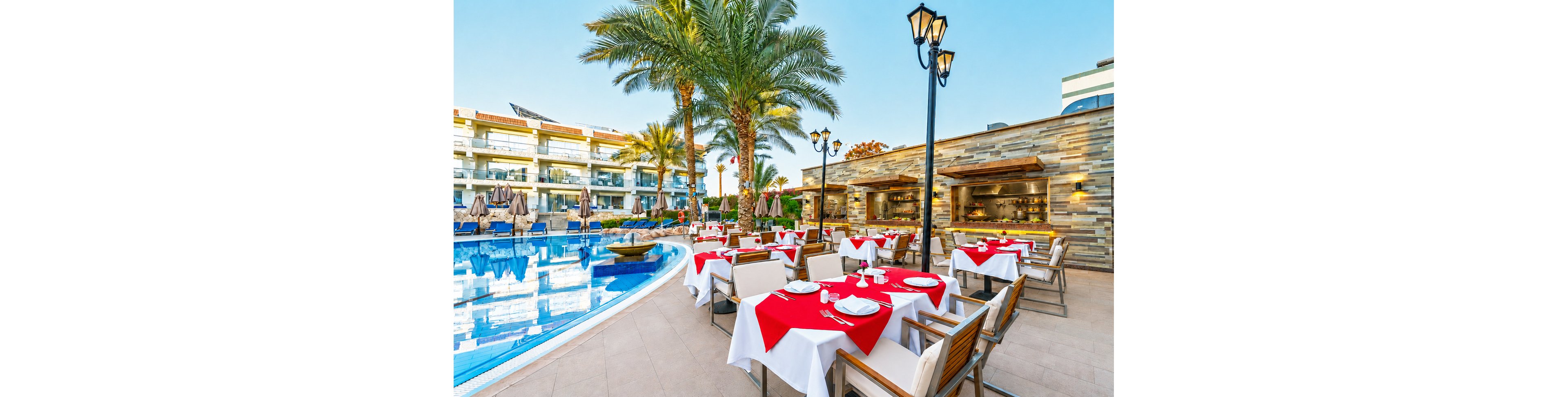 An elegant outdoor restaurant is set beside a curved swimming pool with clear blue water. Tables are neatly arranged with red tablecloths, white napkins, and simple place settings, creating a vibrant dining area. Tall palm trees and modern resort buildings frame the scene, suggesting a warm vacation destination. Soft lighting and open-air architecture contribute to a relaxed, inviting atmosphere for guests.