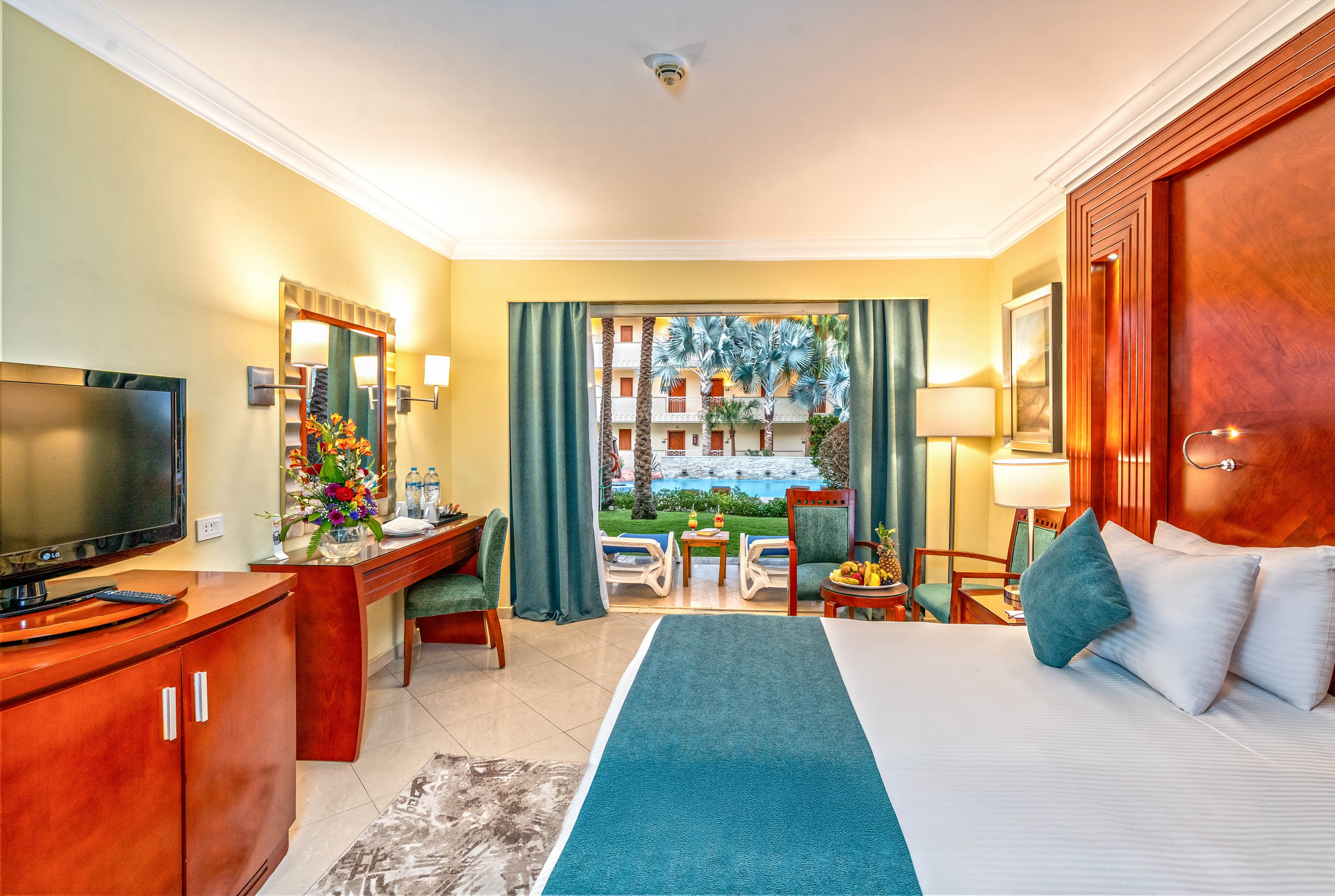 A neatly made hotel room features a large bed with teal accents and multiple pillows. A wooden desk with a flat-screen TV, flowers, and bottled water sits to the side, while a small seating area faces the window. Floor-to-ceiling curtains open to reveal a balcony with chairs overlooking a lush garden and palm trees. Warm lighting and polished wooden furniture create a welcoming, vacation-ready atmosphere.