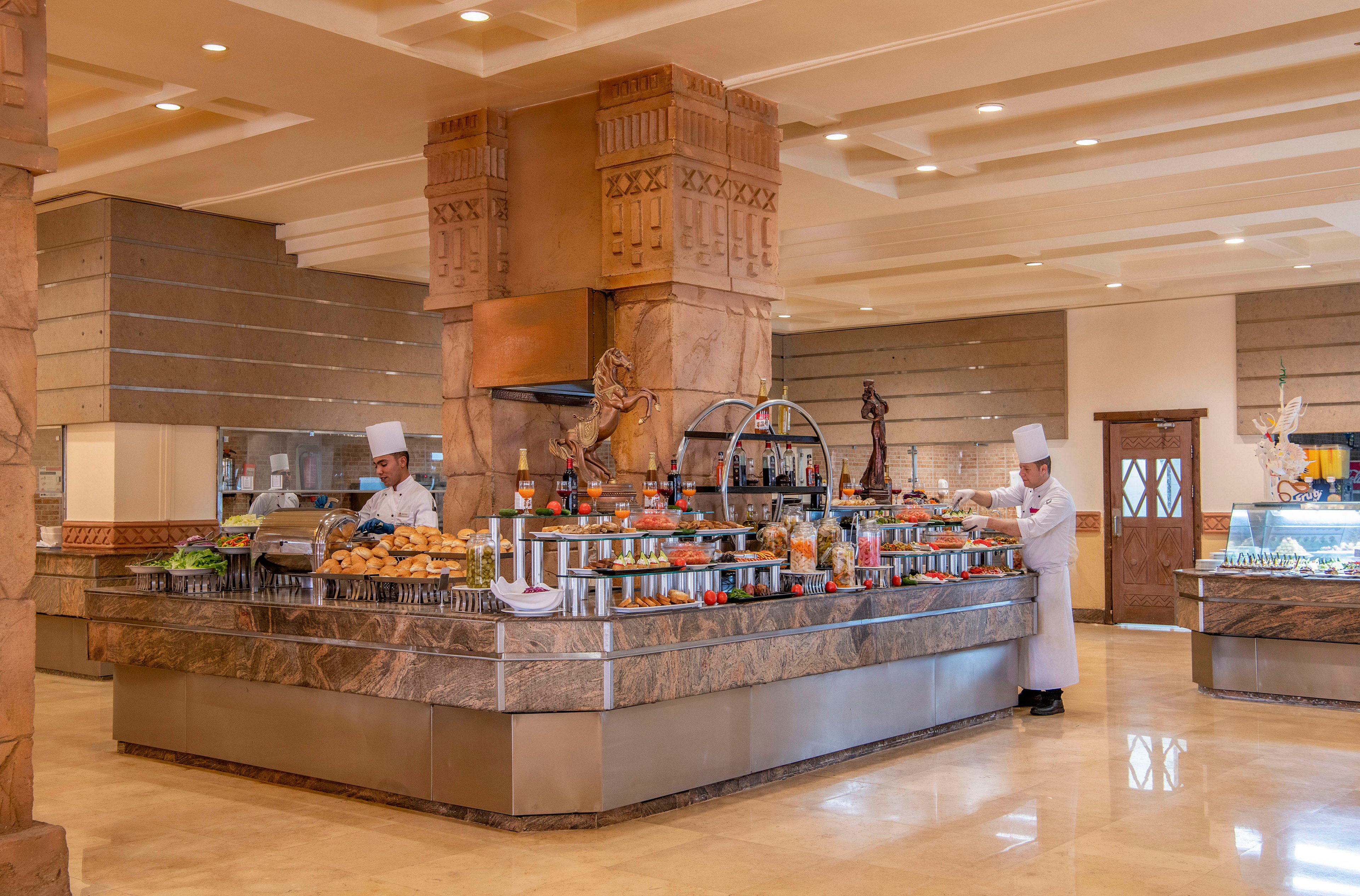 Two chefs in white uniforms are seen preparing and arranging a buffet in a spacious and elegant dining area. The setting features a central food station with a variety of dishes, condiments, and beverages. The decor includes warm tones, marble surfaces, and intricate architectural details, creating a luxurious ambiance.