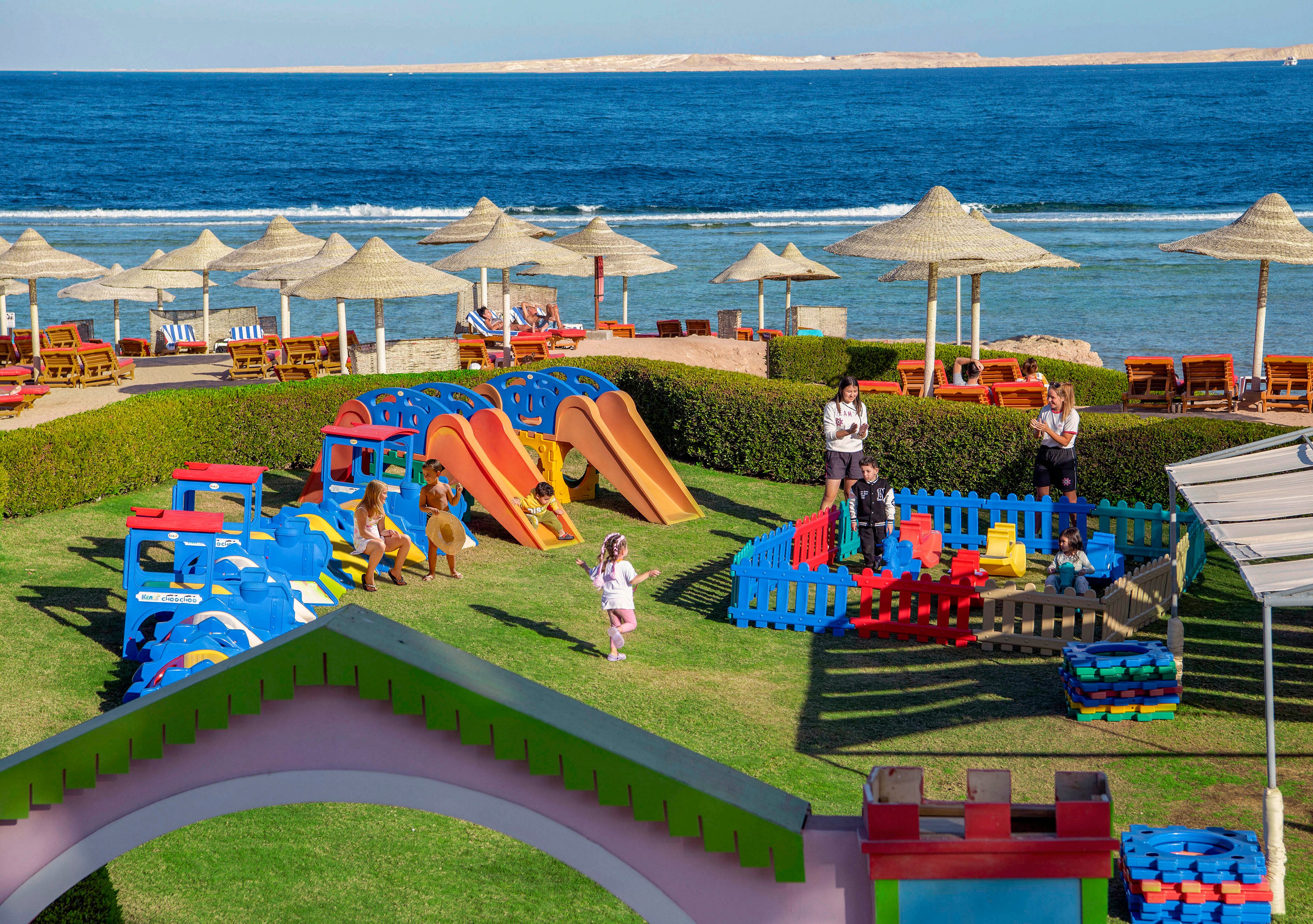 A vibrant children's playground featuring colorful slides, toy trains, and play structures is set against a scenic seaside backdrop. The area is surrounded by green lawns, umbrellas, and lounge chairs, creating a family-friendly environment. Adults and children are seen enjoying the space under clear blue skies.