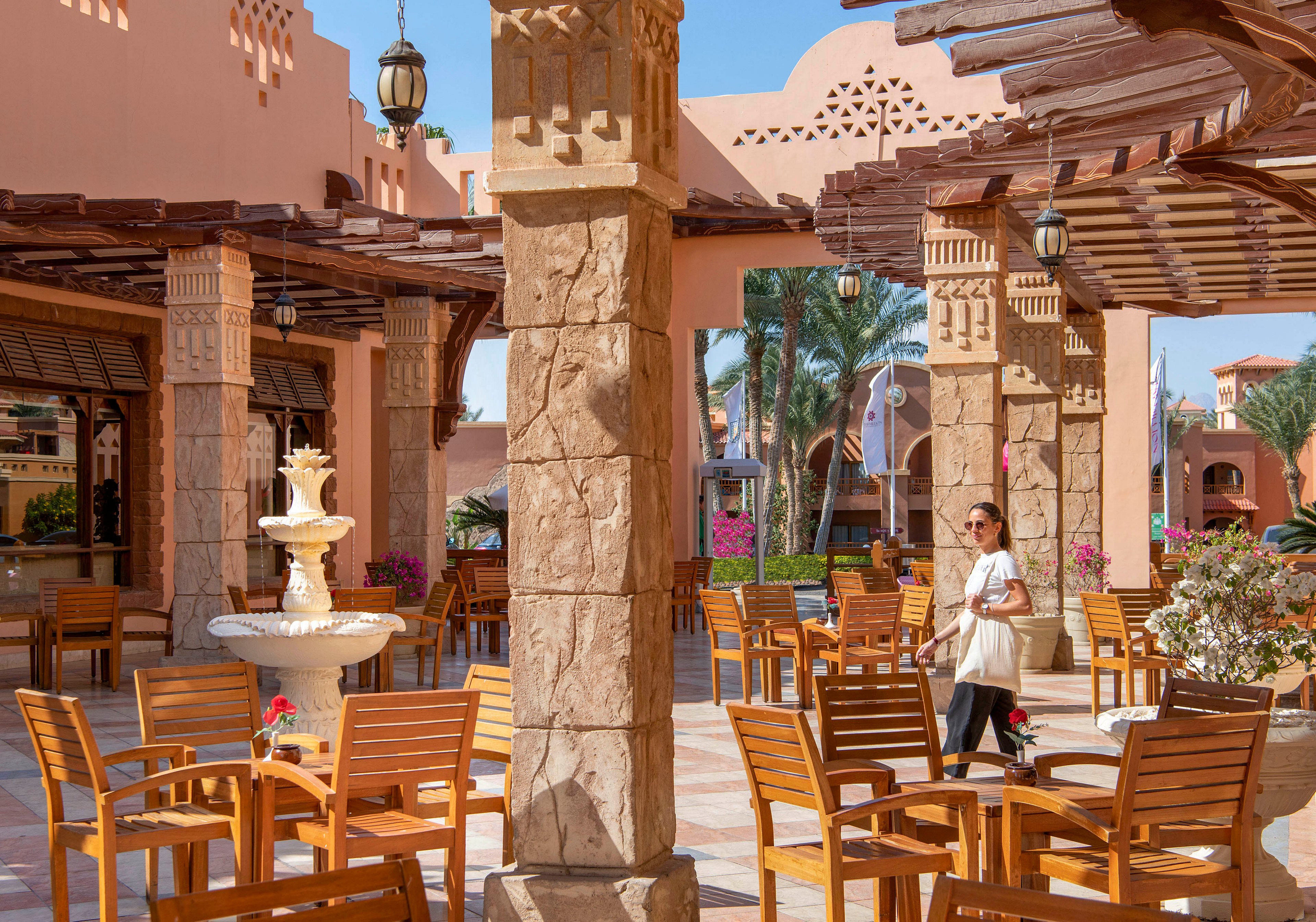 An inviting outdoor cafe setting featuring wooden chairs and tables arranged around a central fountain. The area is shaded by wooden pergolas and surrounded by vibrant pink flowers and palm trees. A person is walking through the space, adding a lively touch to the serene atmosphere.