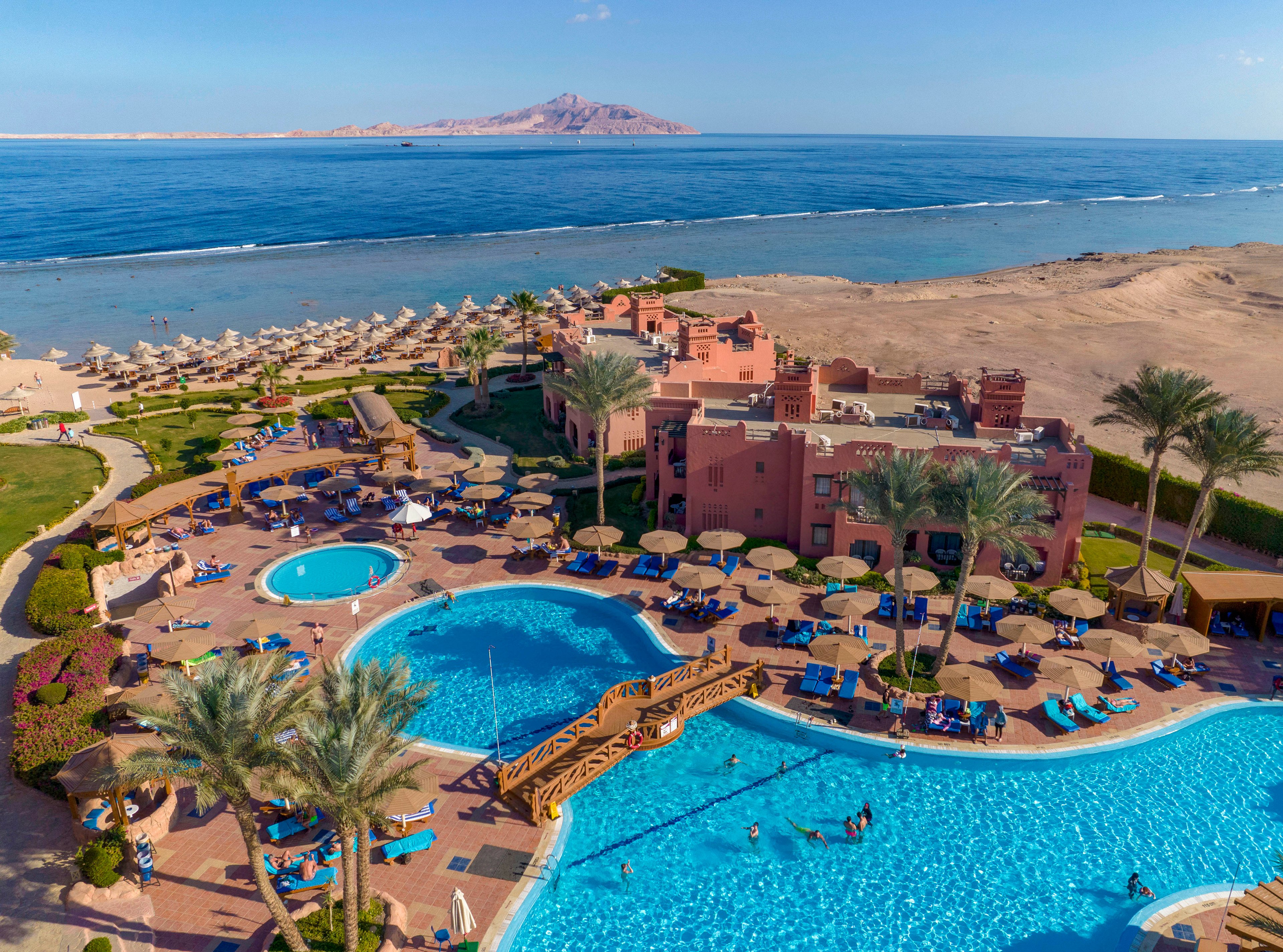 A scenic view of a luxury resort featuring multiple swimming pools, sun loungers, and a wooden bridge. The resort is located by the sea, with a sandy beach and umbrellas lining the shore. In the background, a mountain rises above the horizon, adding to the picturesque setting.