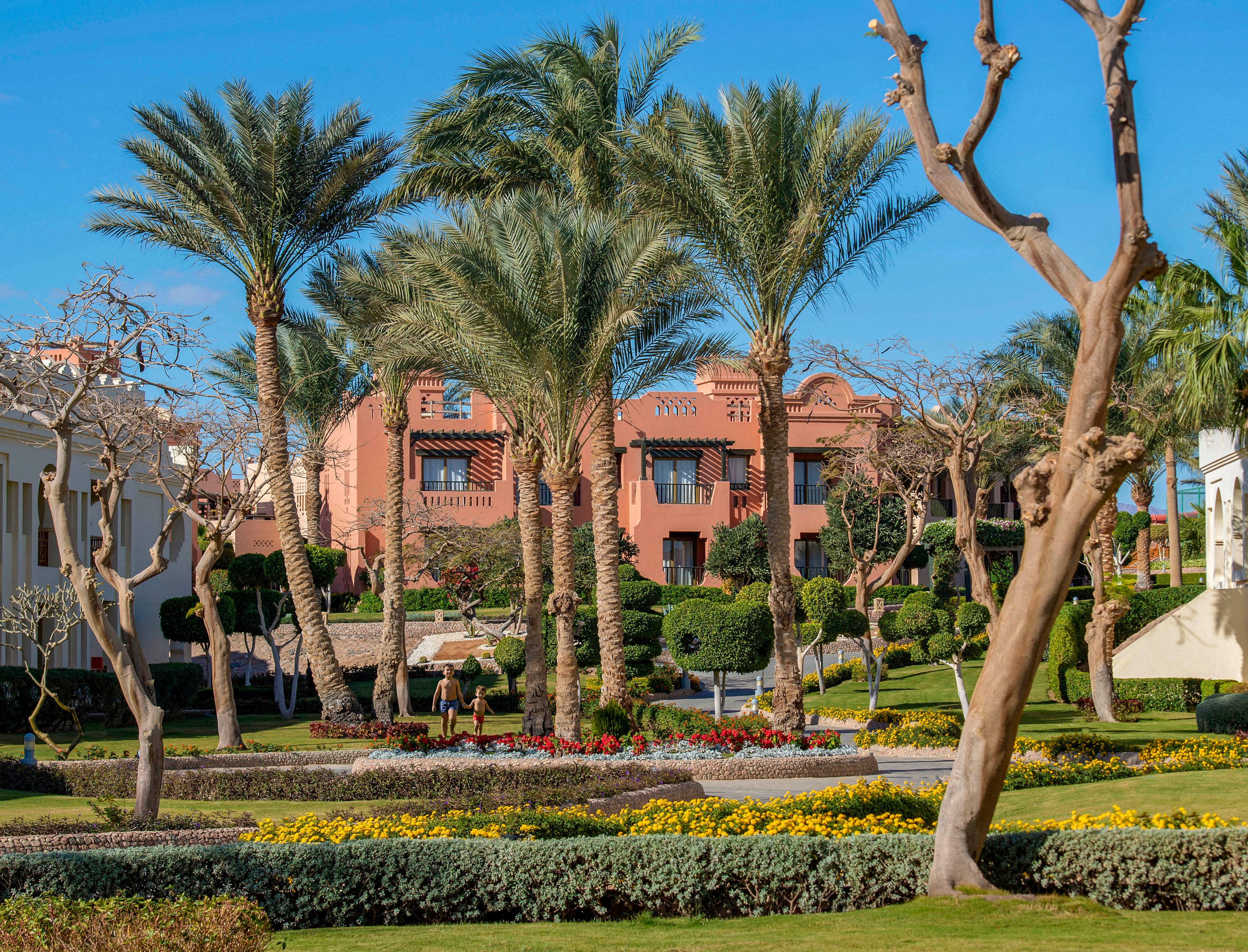 A luxurious villa with terracotta walls is nestled amidst a beautifully landscaped garden featuring palm trees, trimmed hedges, and vibrant flower beds. The setting is sunny with a clear blue sky, evoking a serene and tropical ambiance. The scene showcases a well-maintained outdoor space ideal for relaxation or leisure.