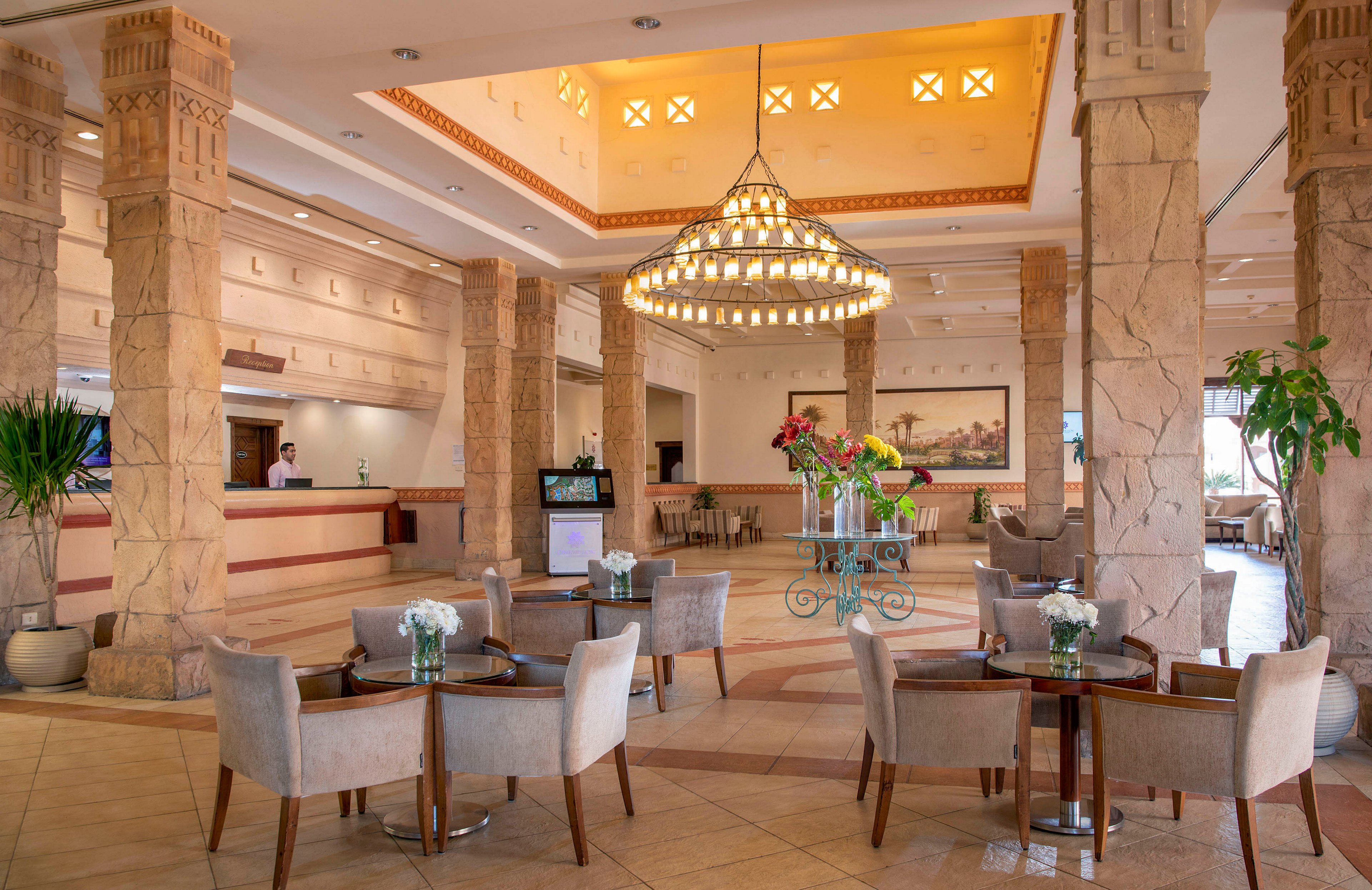 A spacious and well-lit hotel lobby featuring a grand chandelier, stone pillars, and a seating area with small tables and chairs. The decor includes floral arrangements and warm lighting, creating a welcoming atmosphere. A reception desk is visible in the background, along with a wall-mounted painting and indoor plants.