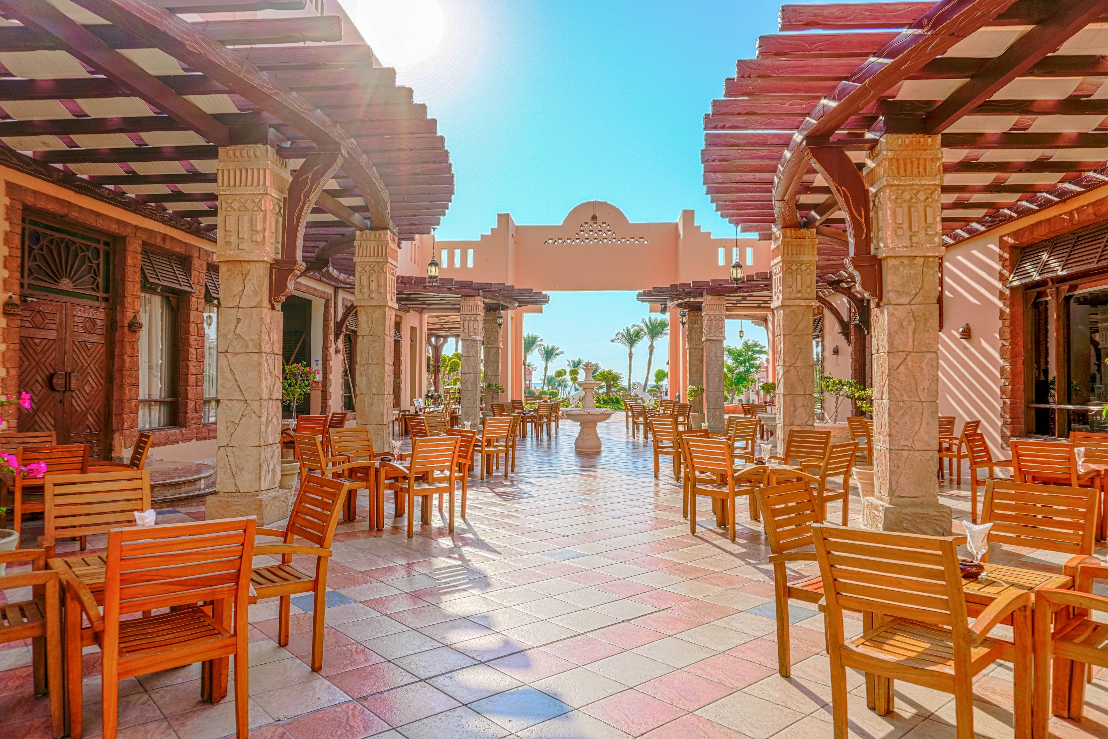 A bright outdoor dining area featuring wooden tables and chairs under pergola-style structures. The setting is sunny with clear blue skies and decorative columns. The space exudes a warm and inviting atmosphere, perfect for relaxation or casual dining.