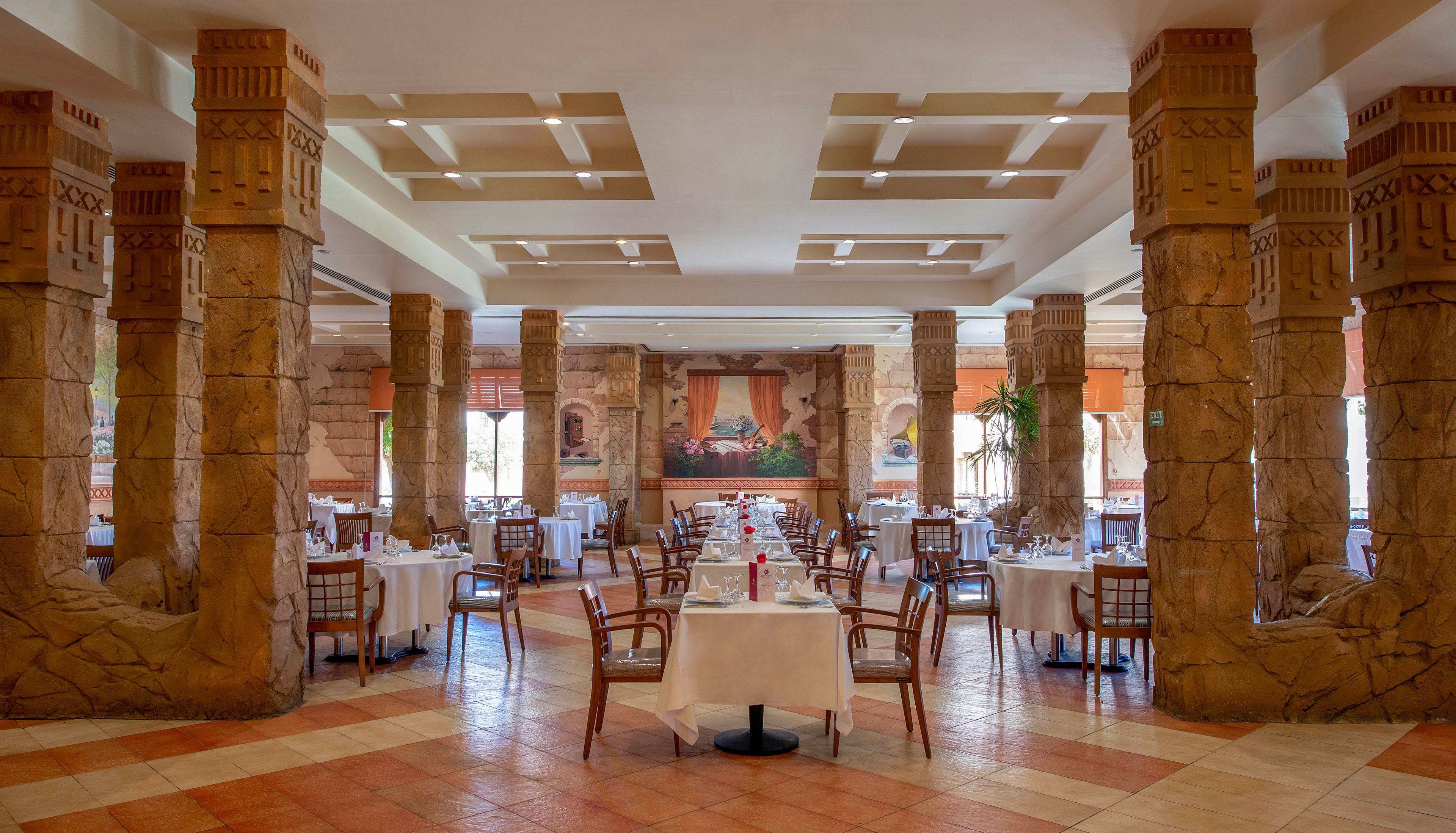 A spacious and elegant restaurant interior featuring intricately designed stone columns and neatly arranged dining tables with white tablecloths. The warm lighting and earthy tones create a welcoming ambiance. Large windows and wall art enhance the aesthetic appeal of the space.
