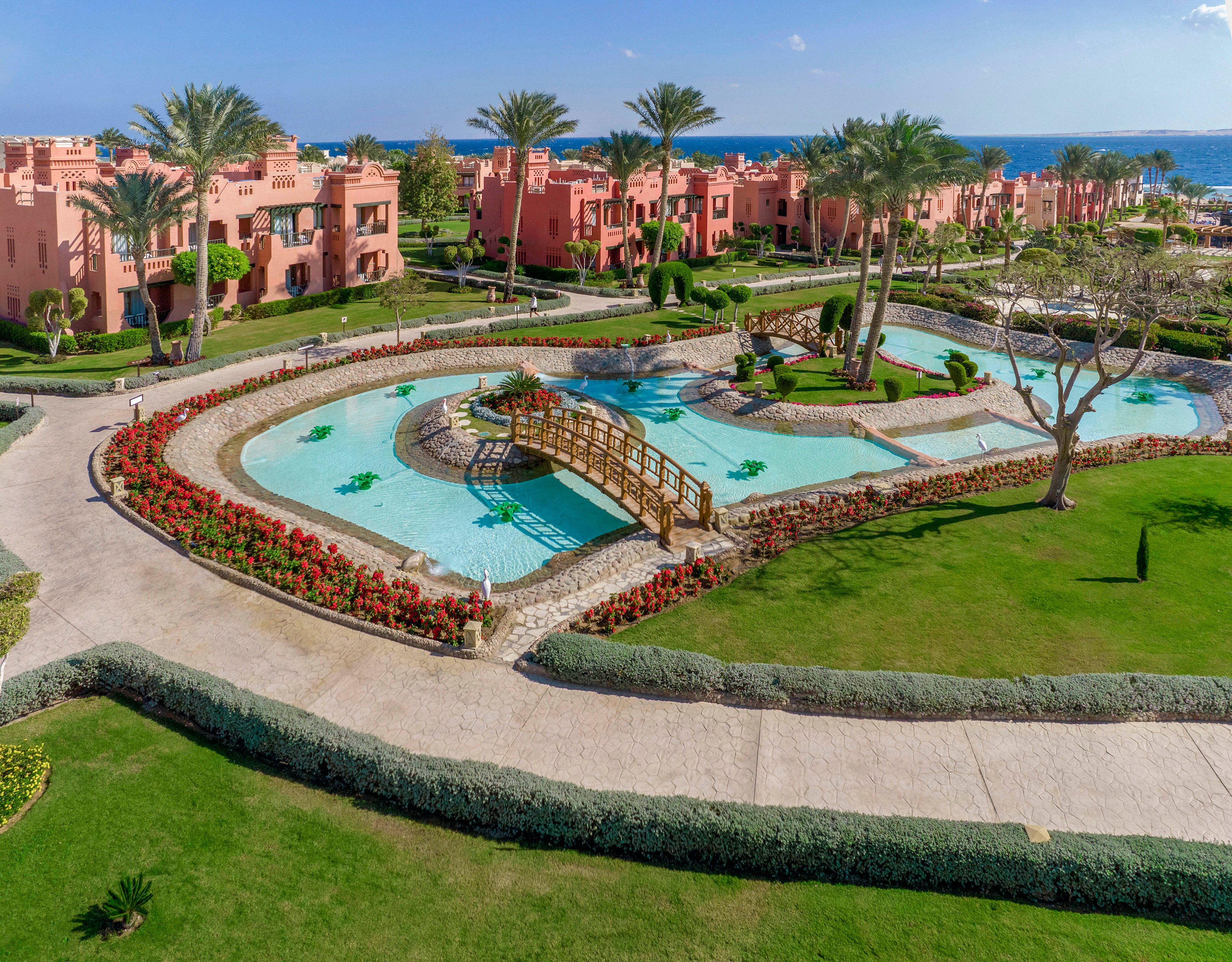 A scenic view of a luxury resort featuring lush landscaped gardens, turquoise pools, and a wooden bridge. The setting includes vibrant red flowers, palm trees, and pink villa-style buildings. The backdrop showcases a clear blue sky and the sea in the distance, creating a serene and tropical atmosphere.