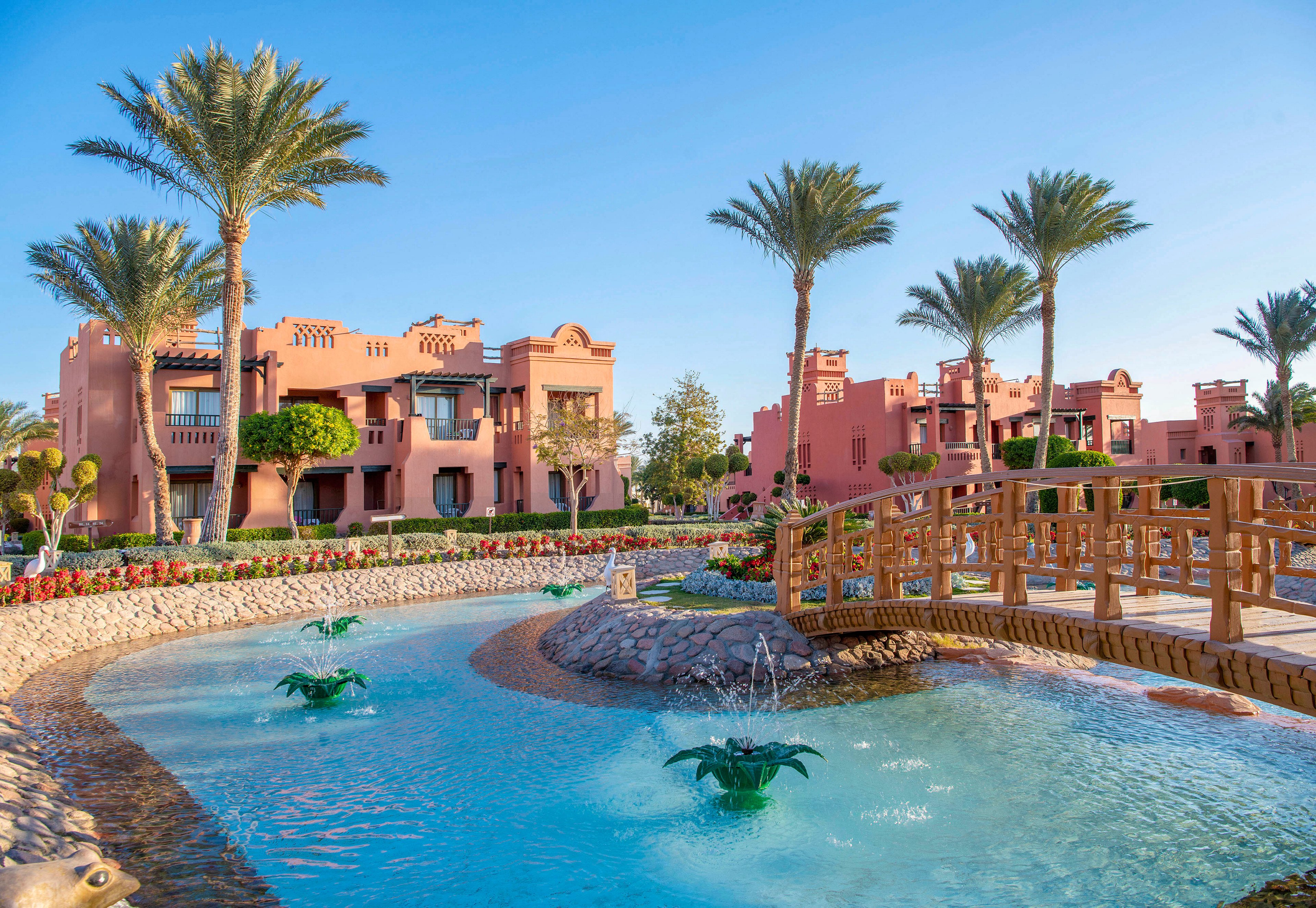 A scenic view of a luxury resort featuring pink stucco buildings surrounded by lush palm trees. A wooden bridge spans over a clear blue pool with decorative fountains. The setting is bright and sunny, evoking a tranquil and tropical atmosphere.
