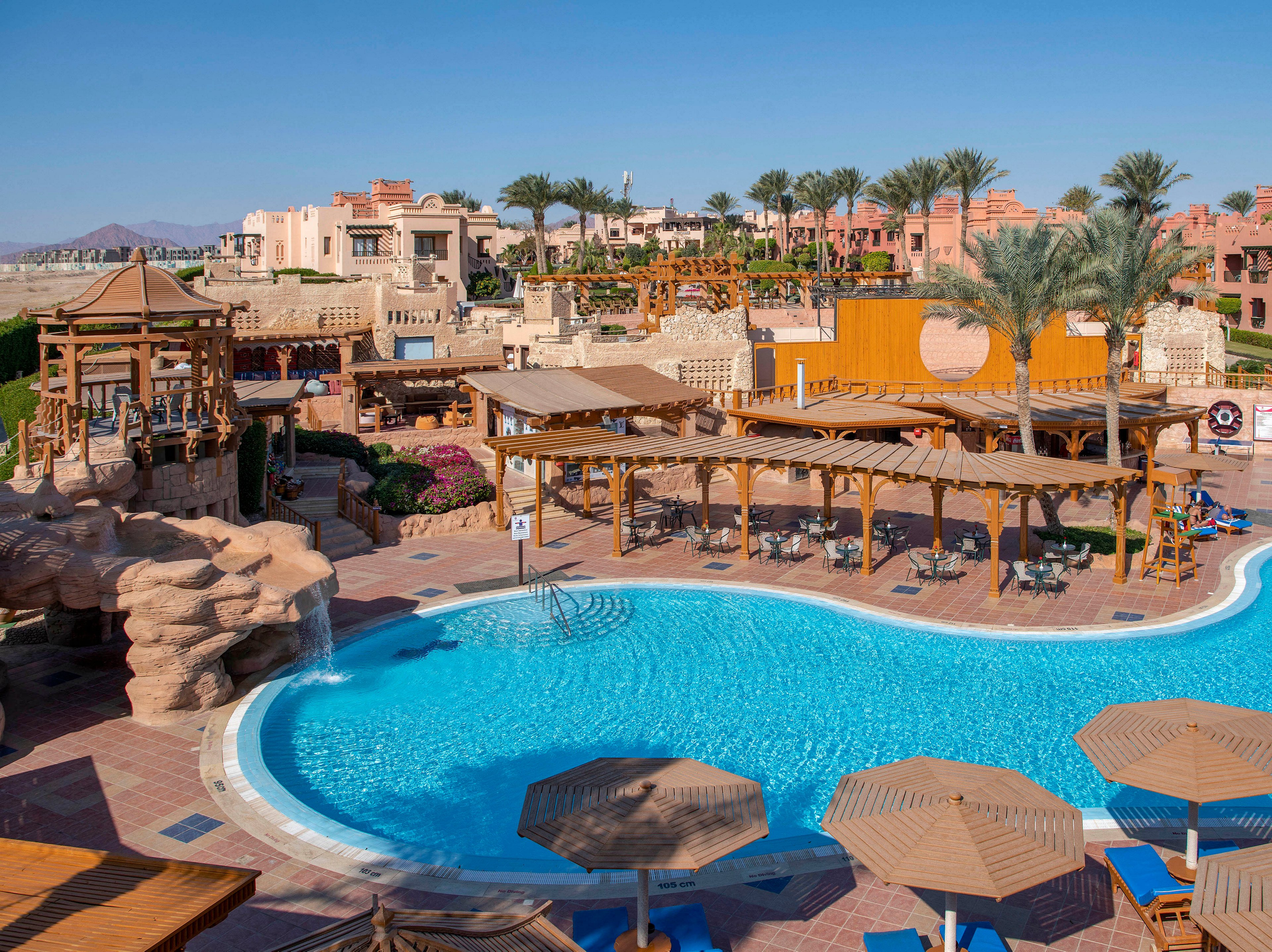A vibrant outdoor pool surrounded by wooden pergolas and palm trees in a luxury resort setting. The scene features clear blue water, sun loungers, and a backdrop of elegant architecture. The sunny weather enhances the inviting atmosphere of the resort.