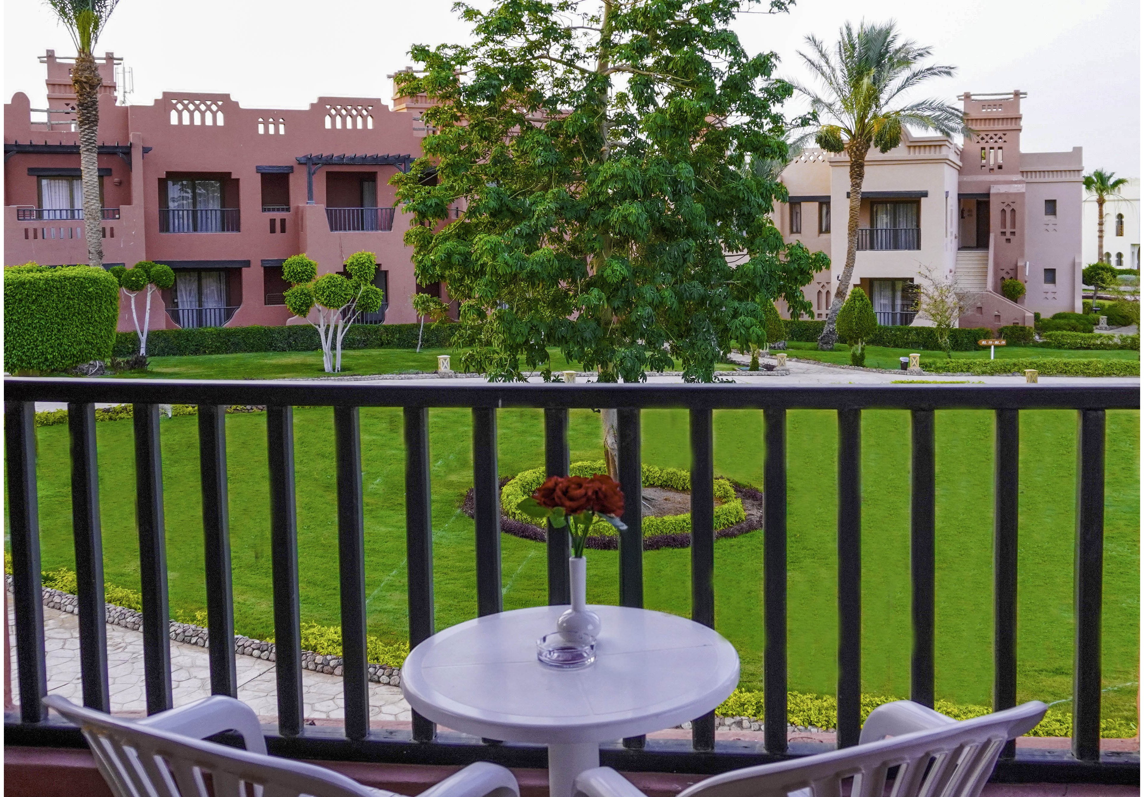A cozy outdoor seating area with a small table and two chairs overlooks a well-maintained garden. The scene features vibrant green grass, neatly trimmed trees, and a backdrop of elegant, villa-style buildings. Two glasses with colorful drinks and a flower arrangement add a touch of charm to the setting.