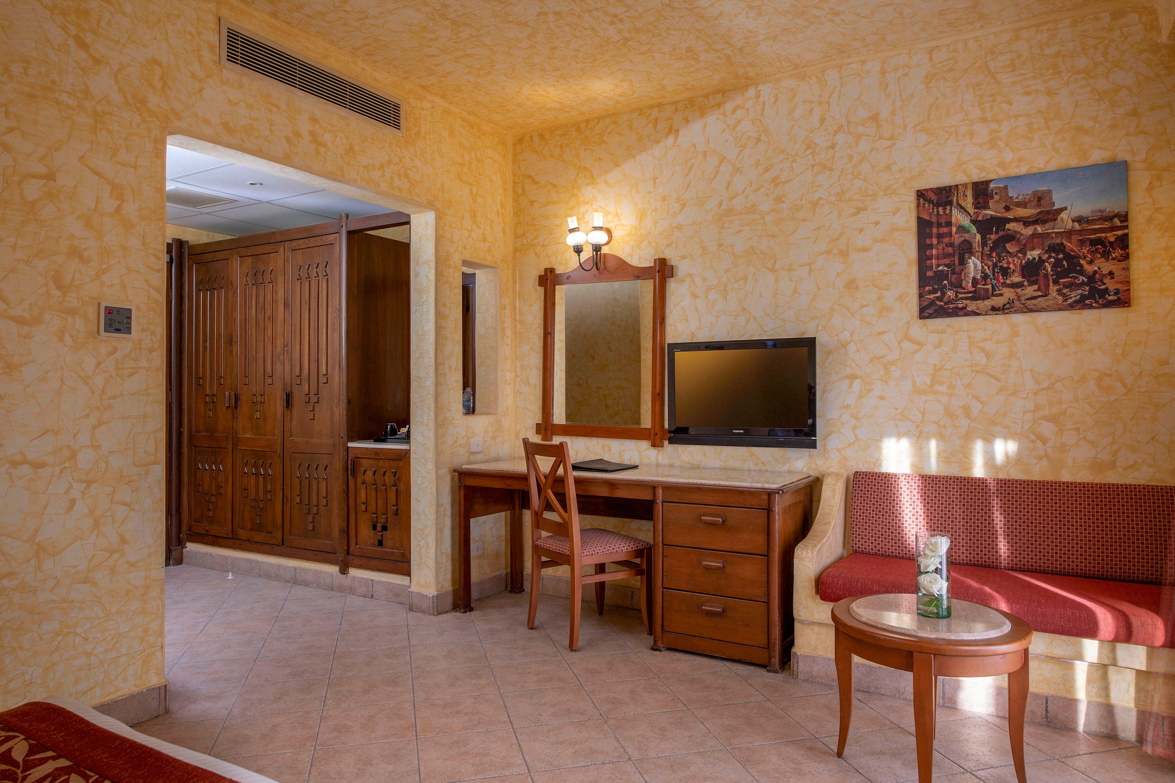 A well-lit hotel room featuring warm yellow textured walls and tiled flooring. The space includes a wooden desk with a mirror, a television, and a seating area with a red cushioned sofa and round table. A wardrobe and additional furniture complete the inviting setup.