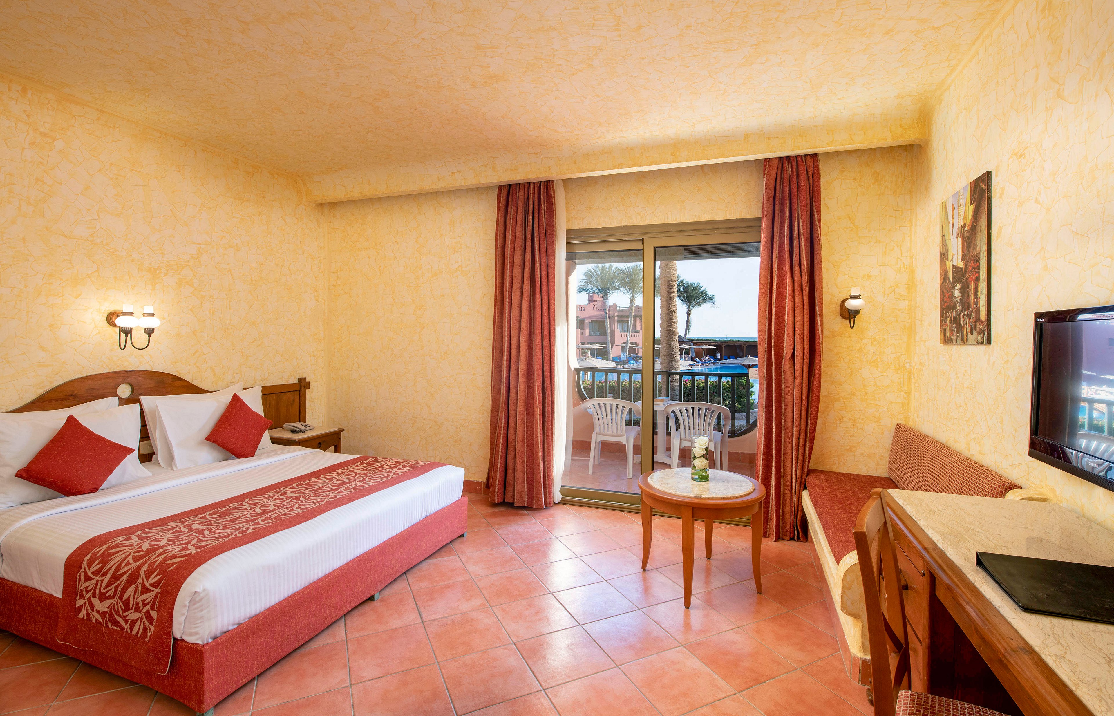 A warmly lit hotel room featuring a double bed with red and white bedding, a small table with chairs, and a balcony overlooking an outdoor area. The room has a flat-screen TV, wooden furniture, and terracotta tile flooring. The walls are painted in a textured yellow finish, creating a cozy ambiance.