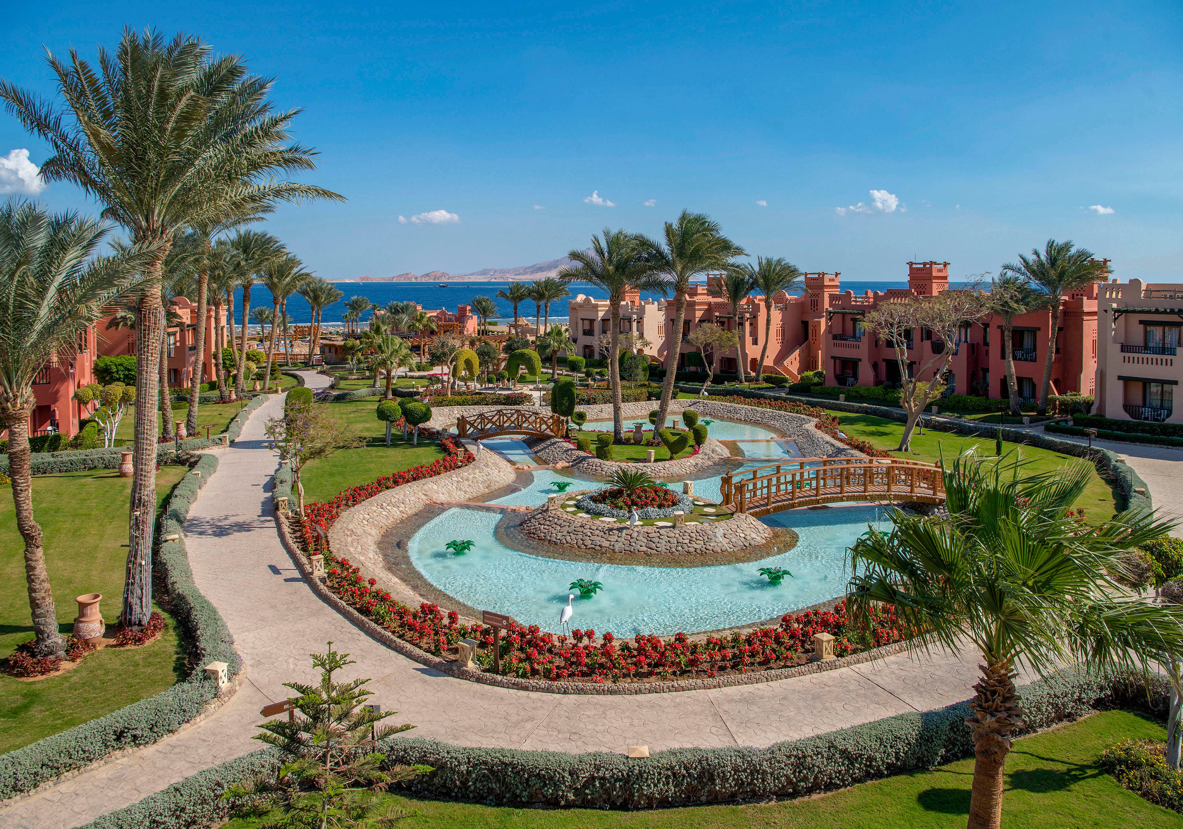 A beautifully landscaped resort garden featuring a winding water feature surrounded by vibrant flowers and palm trees. The setting includes elegant pathways, wooden bridges, and a backdrop of red-toned buildings. The clear blue sky and distant sea view enhance the serene and tropical atmosphere.