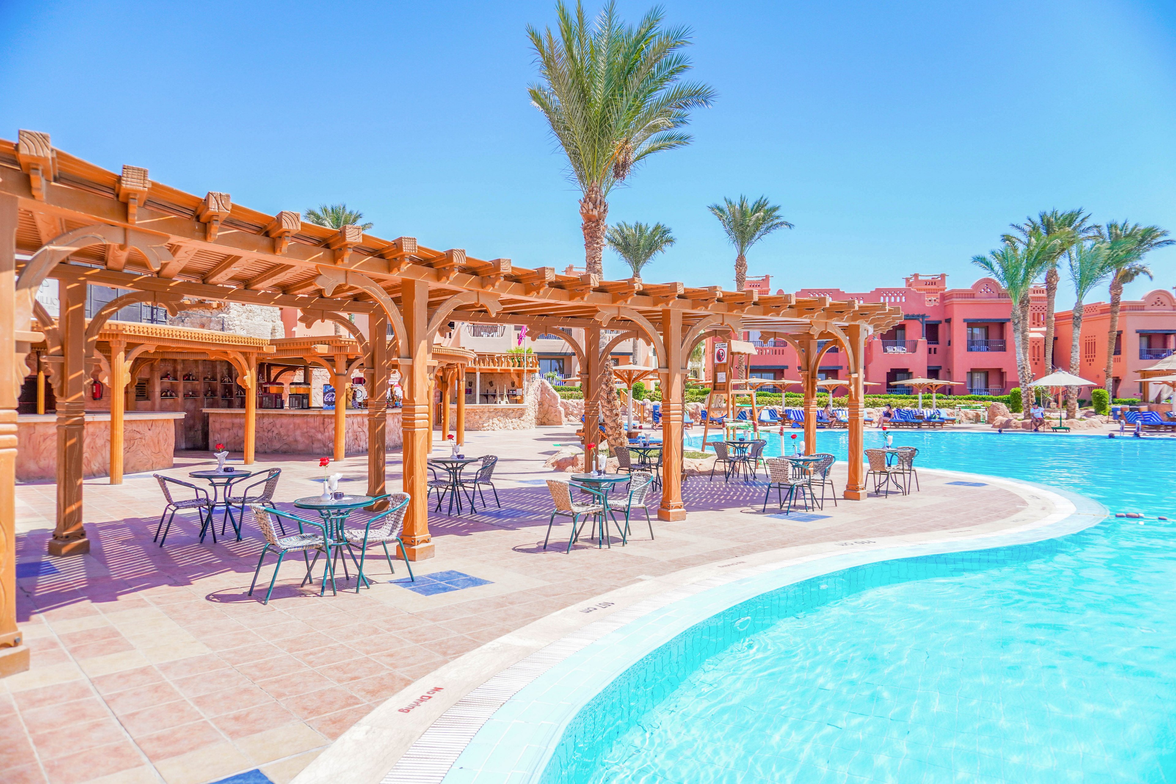 A serene resort setting featuring a large swimming pool surrounded by wooden pergolas and outdoor seating. Palm trees and vibrant architecture create a tropical ambiance under a clear blue sky. The area is designed for relaxation and leisure, with tables and chairs arranged for guests.