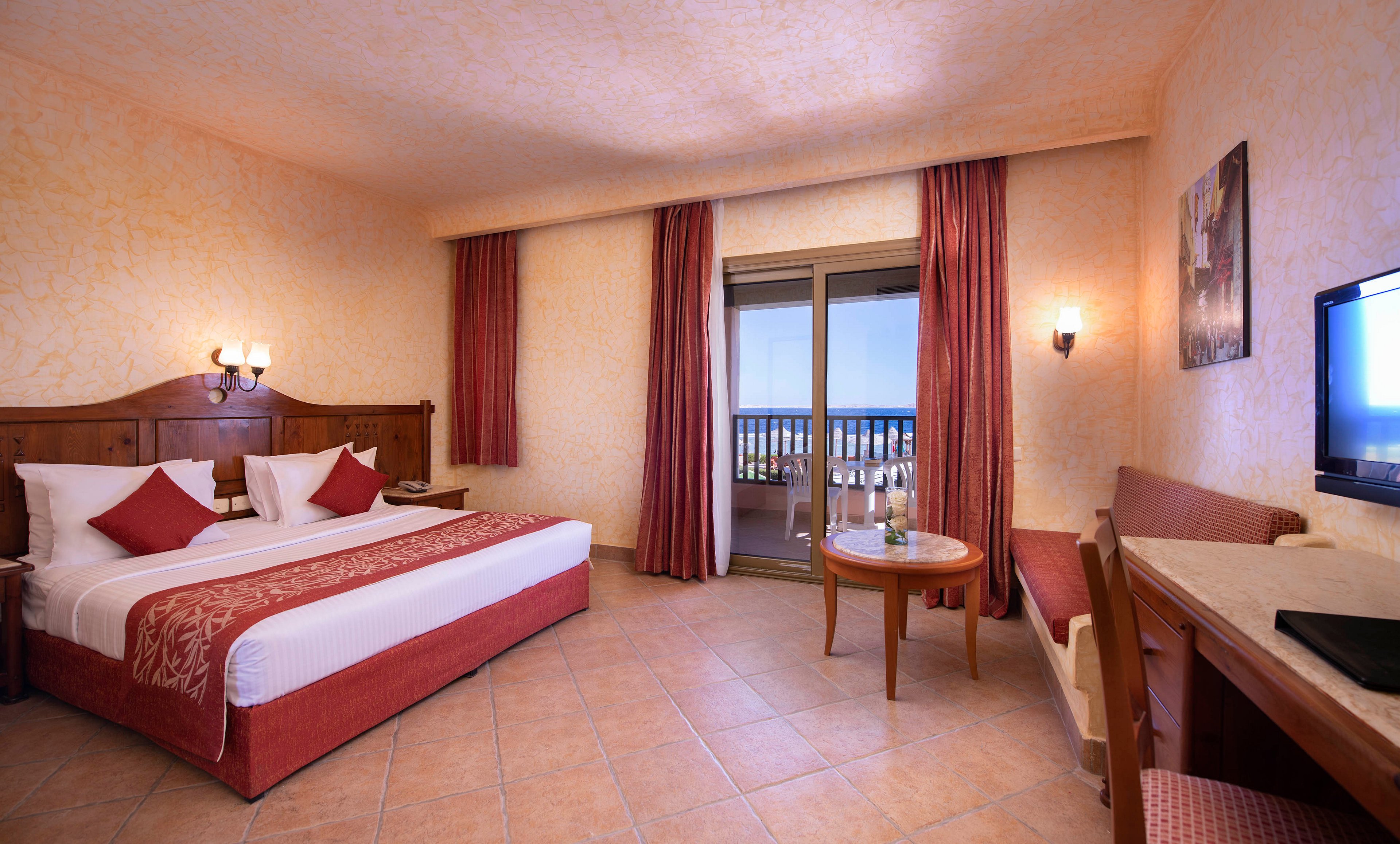 A warm and inviting hotel room featuring a large bed with red and white bedding, a seating area, and a desk. The room is decorated in earthy tones with matching curtains and walls. A balcony with a scenic view is visible, adding to the relaxing ambiance.