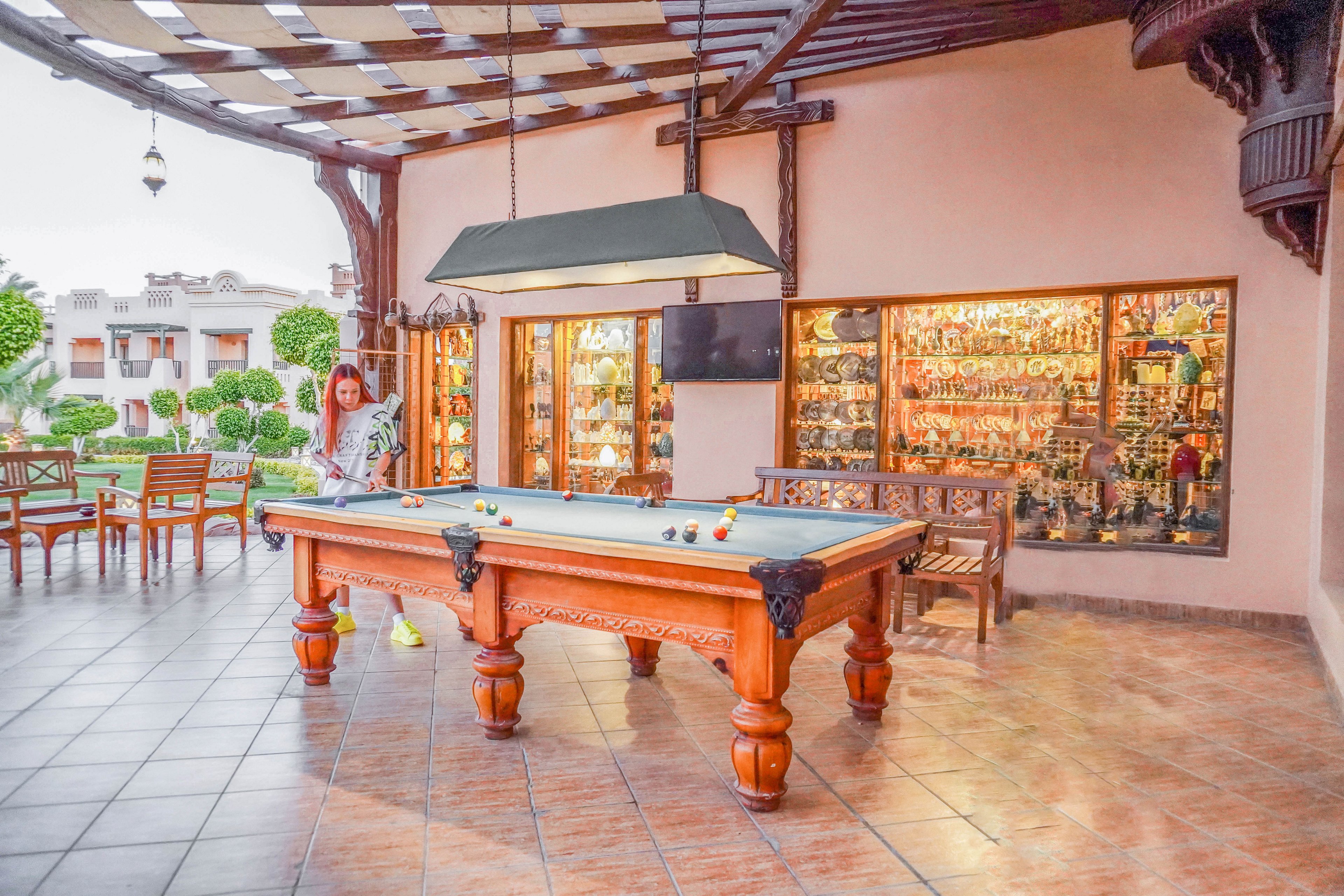 An elegant wooden billiards table is set in a covered outdoor patio with tiled flooring. A woman stands near the table, preparing for a game. The background features a display of decorative items and a seating area, with lush greenery visible outside.