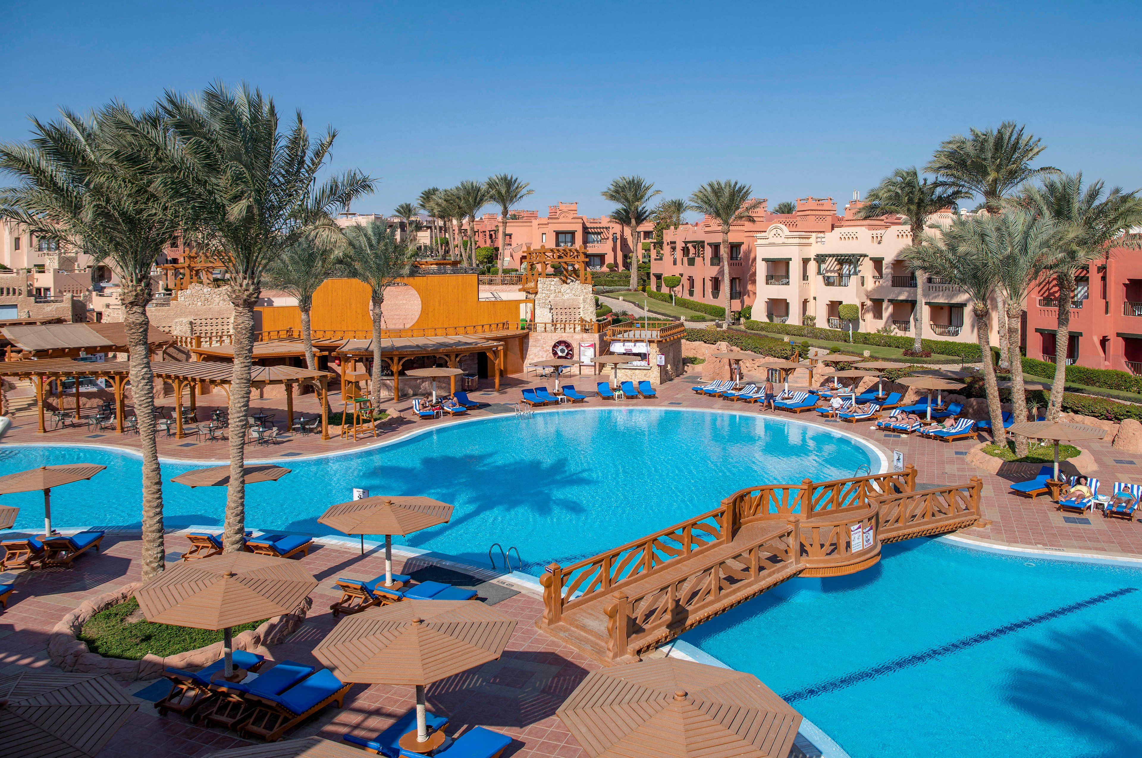 A luxurious resort pool surrounded by palm trees and sun loungers under umbrellas. The setting features a wooden bridge over the pool and vibrant architecture in the background. The clear blue sky enhances the tropical and relaxing atmosphere.