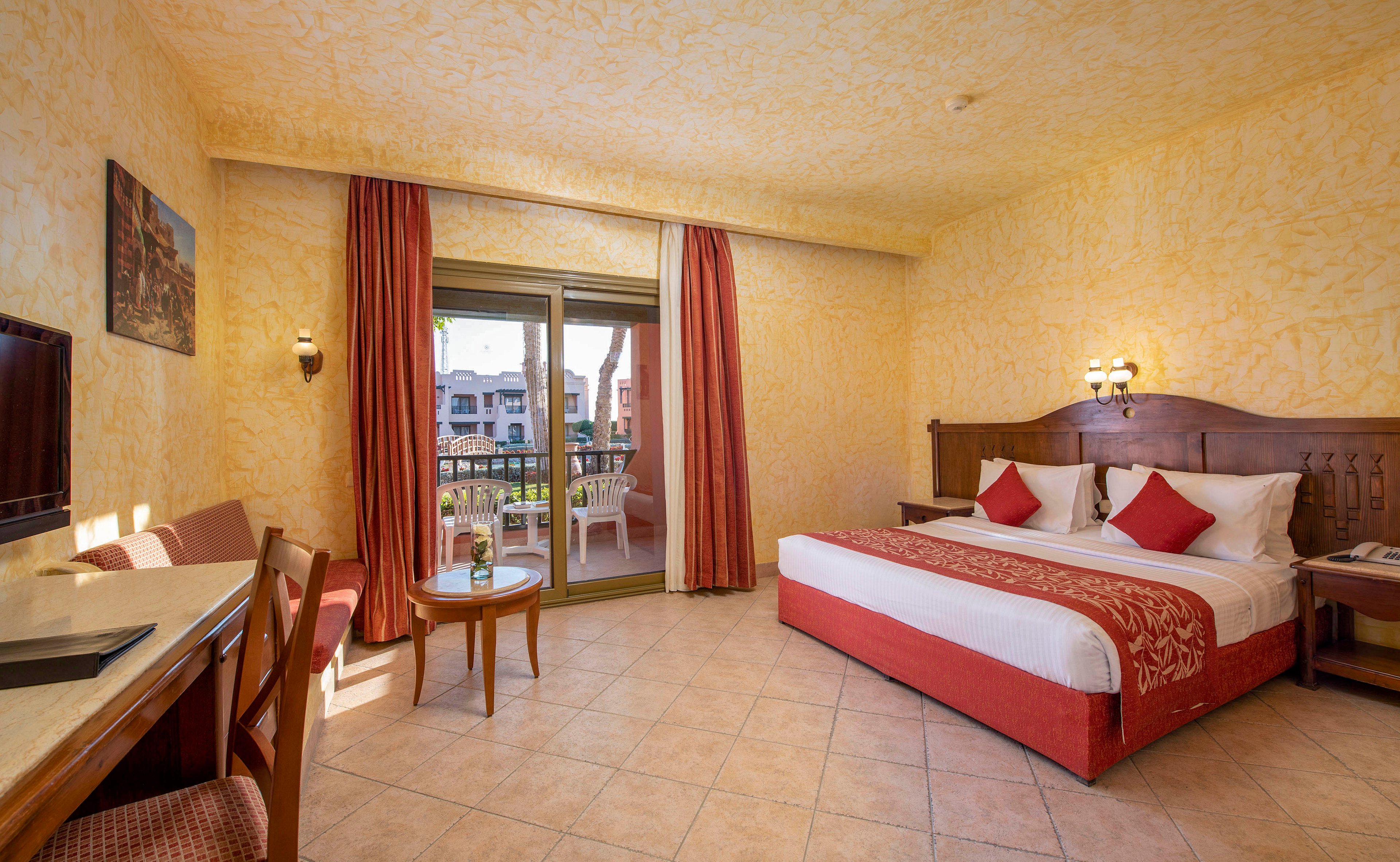 A warmly lit hotel room featuring a large bed with red accents, a small seating area, and a desk. The room opens to a balcony with a view of a sunny outdoor area. The walls have a textured yellow finish, complementing the tiled floor and wooden furniture.
