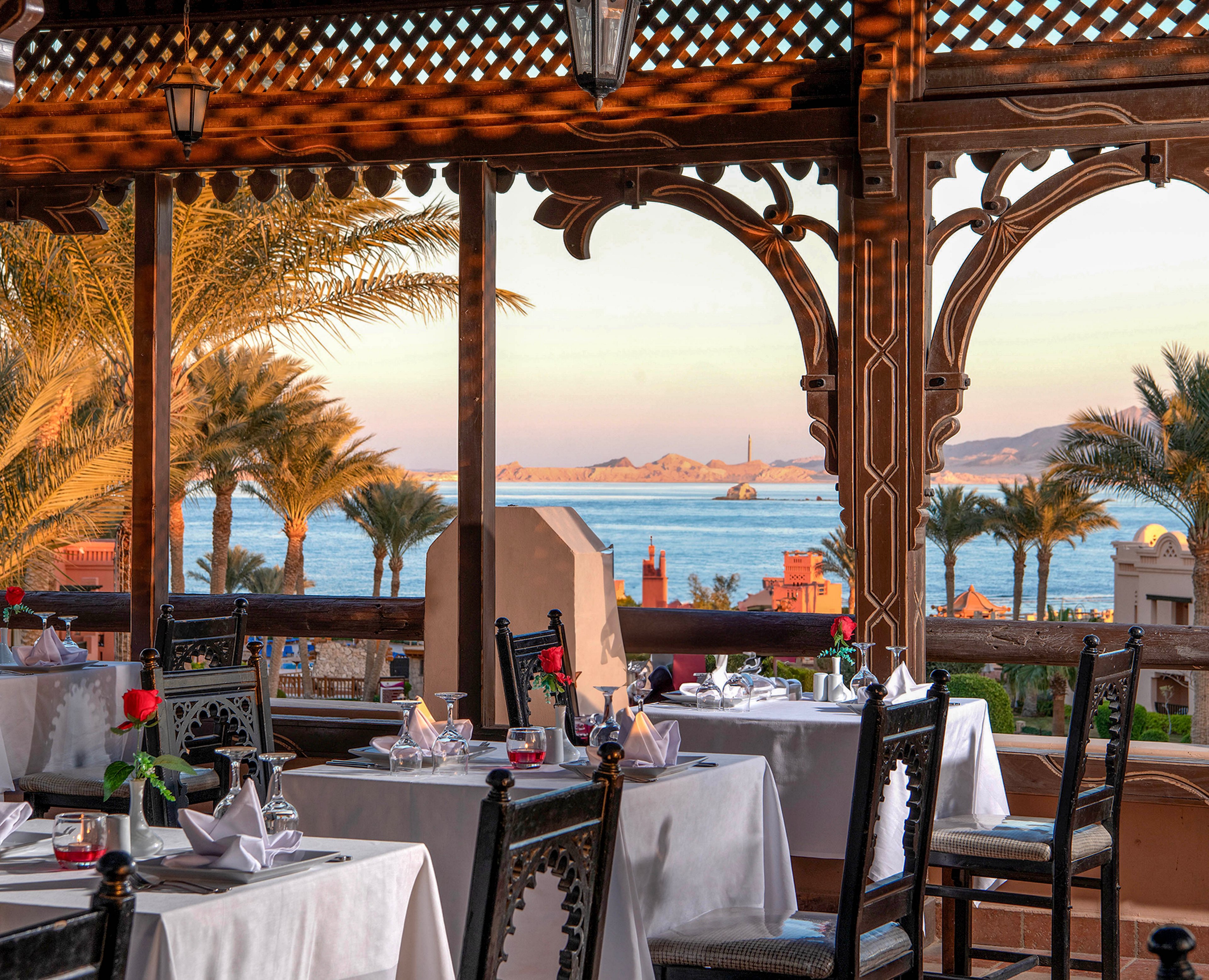 An elegant outdoor dining area with neatly set tables and chairs overlooks a serene ocean view. The setting features intricate wooden decor and lush palm trees, creating a warm and inviting atmosphere. The backdrop includes a calm sea, distant islands, and a soft sunset glow.