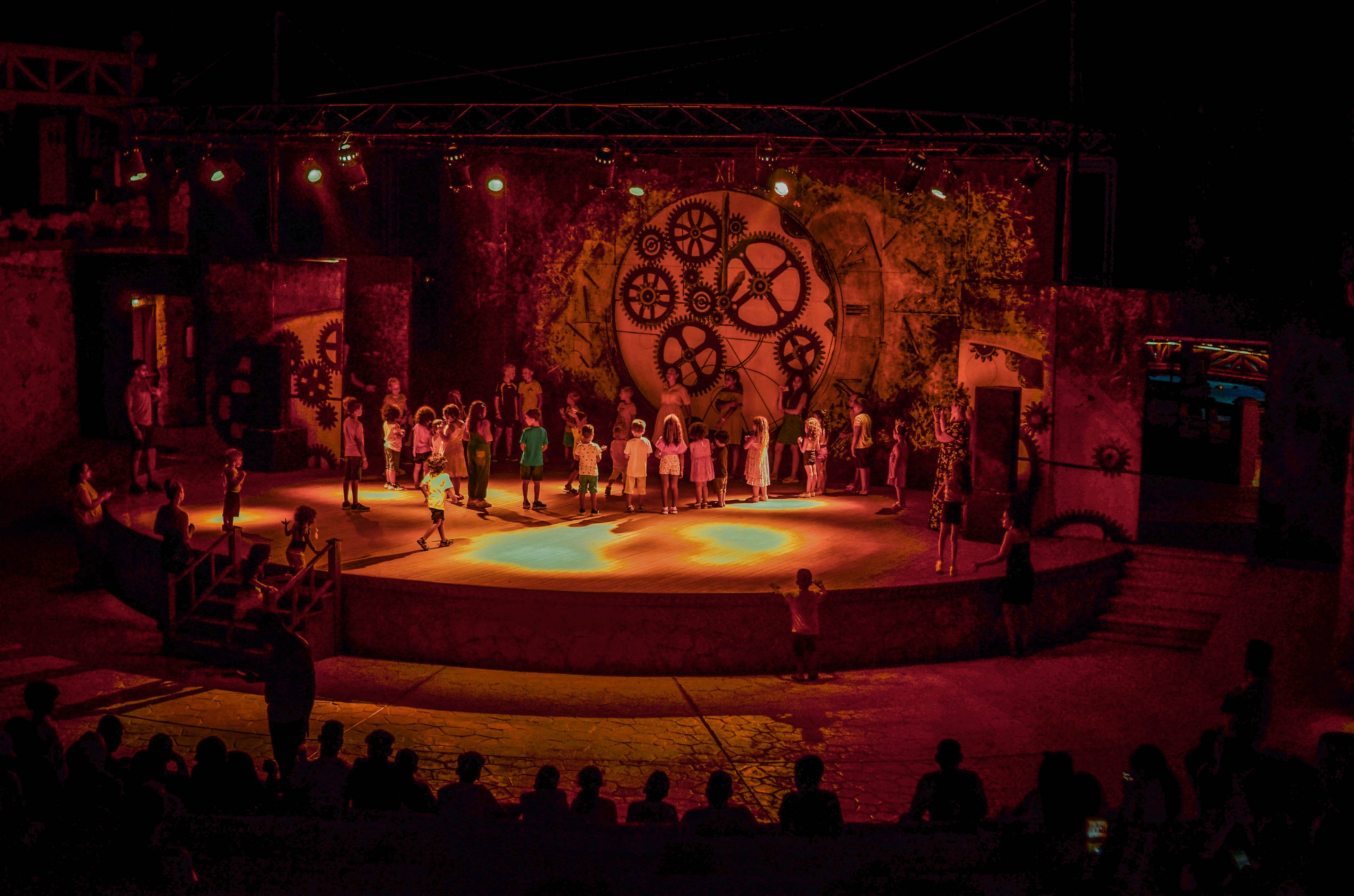 A group of performers stands on a brightly lit stage during a theatrical production. The backdrop features large mechanical gears, adding an industrial aesthetic to the scene. The audience is visible in silhouette, enhancing the dramatic atmosphere of the nighttime setting.