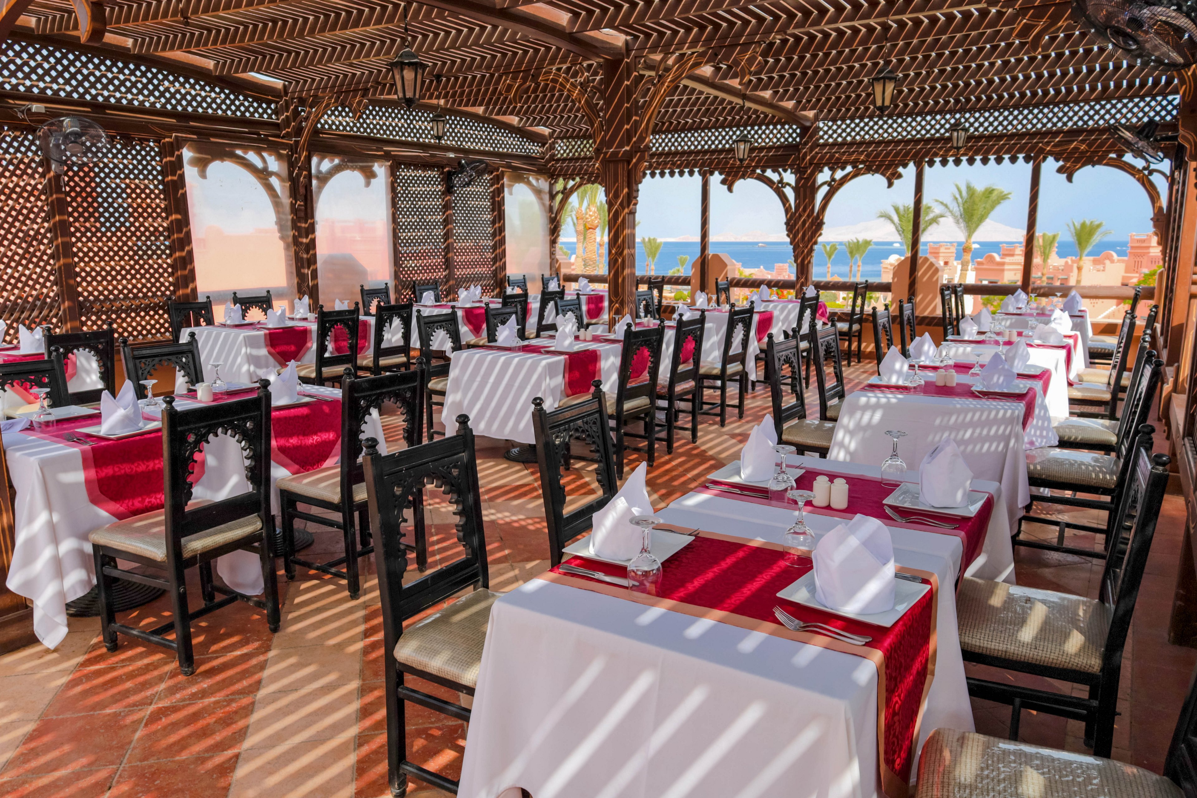 An open-air restaurant featuring neatly arranged tables with white tablecloths and red accents. The setting includes wooden latticework and arches, offering a scenic view of palm trees and the ocean. The atmosphere is sophisticated and inviting, ideal for dining in a serene environment.